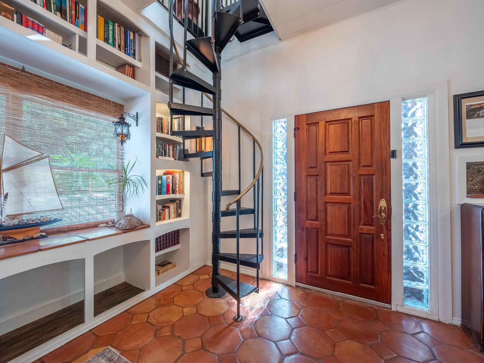 Interior view of a home with a wooden front door, brick-tile flooring, a built-in white bookshelf filled with books, a window with bamboo blinds, and a metal spiral staircase.