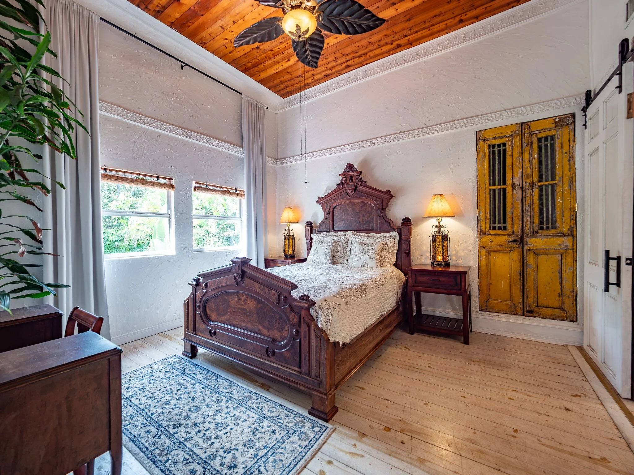 A bedroom with a wooden ceiling featuring a ceiling fan with black leaves and a light, white textured walls, two windows with blinds, a wooden bed with intricate carvings, two yellow lamps on nightstands, a yellow distressed door, and a blue patterne