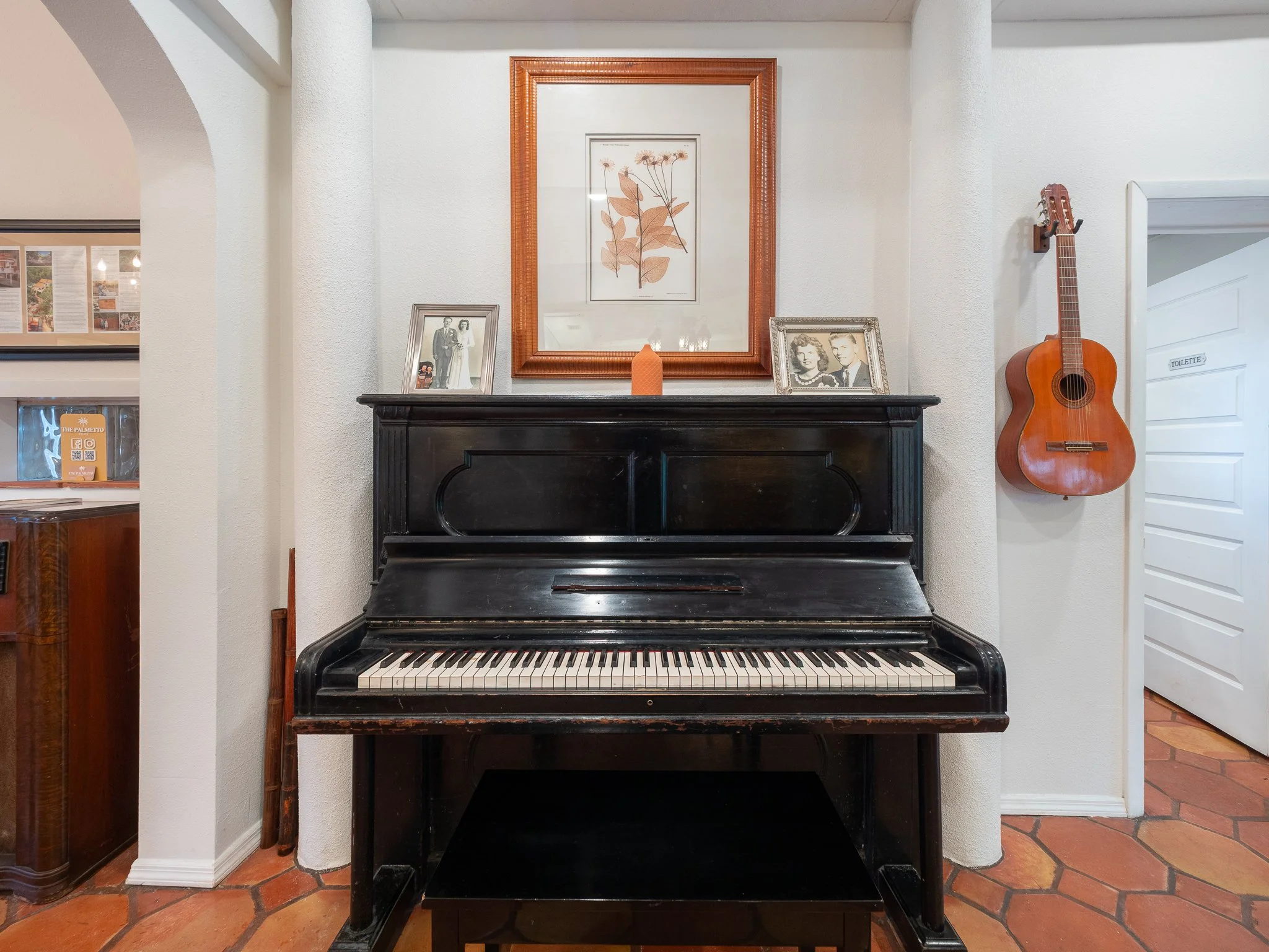A black upright piano with a black finish, placed against a white wall, decorated with two framed photographs, a small orange vase, and a large framed botanical print centered above it. To the right, a wooden guitar hangs on the wall. To the left, pa