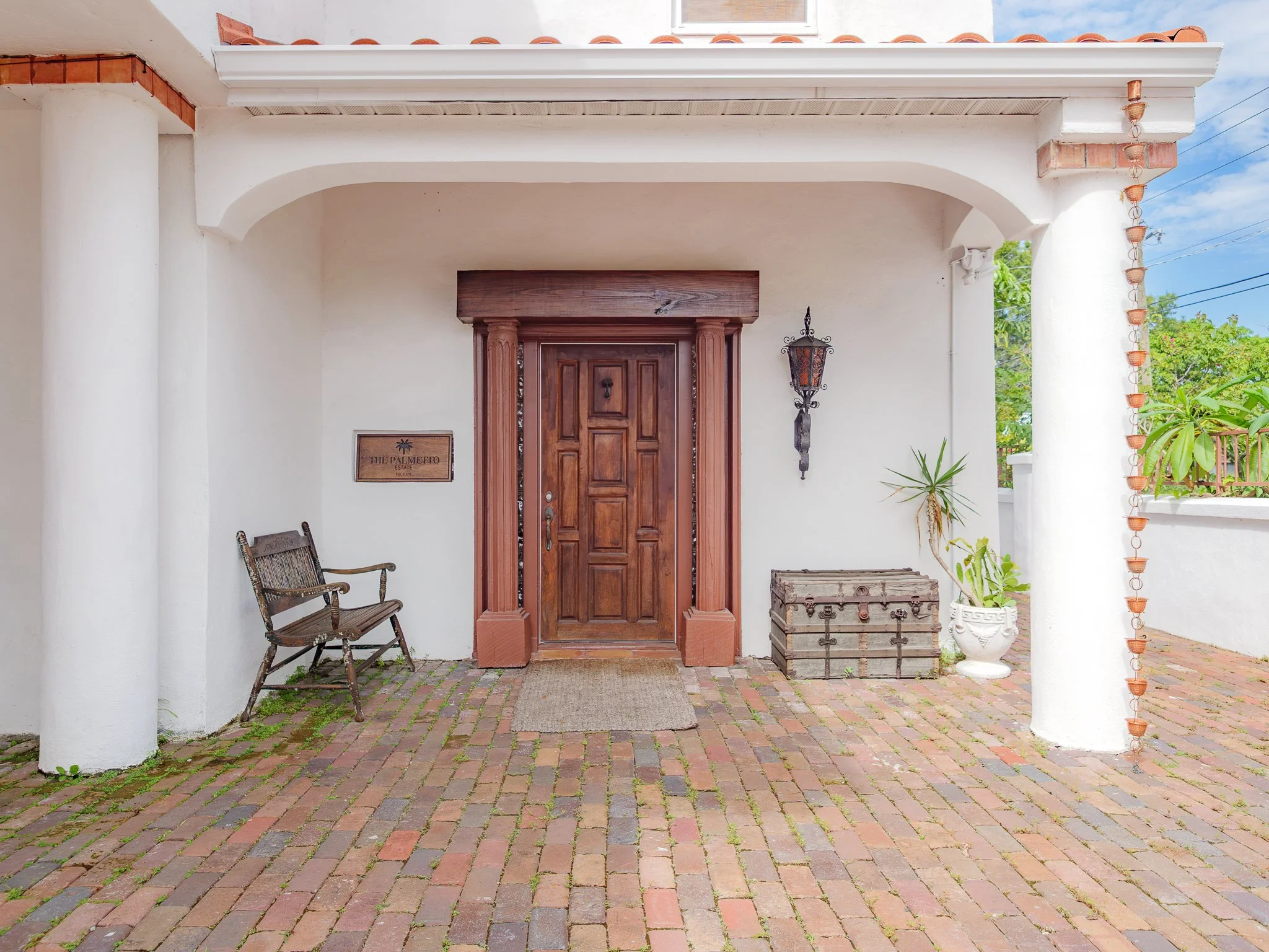 Front porch with wooden door, white stucco walls, brick flooring, patio furniture, potted plants, and wrought iron Spanish light fixture.
