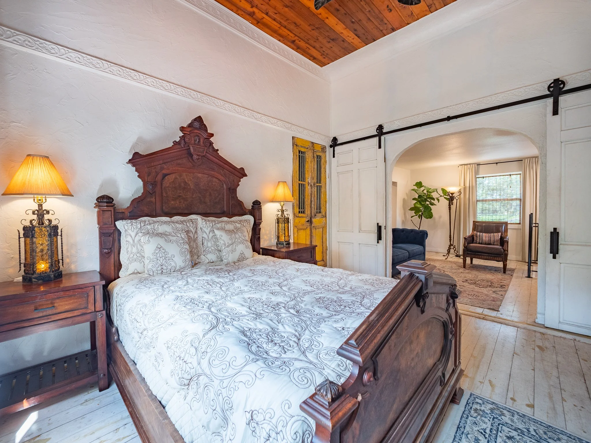 A bedroom with a wooden bed frame and white bedding with embroidered patterns. There are matching nightstands with lamps on either side of the bed. The room has rustic decor, with a wooden ceiling and a mixture of vintage furniture, including a blue 