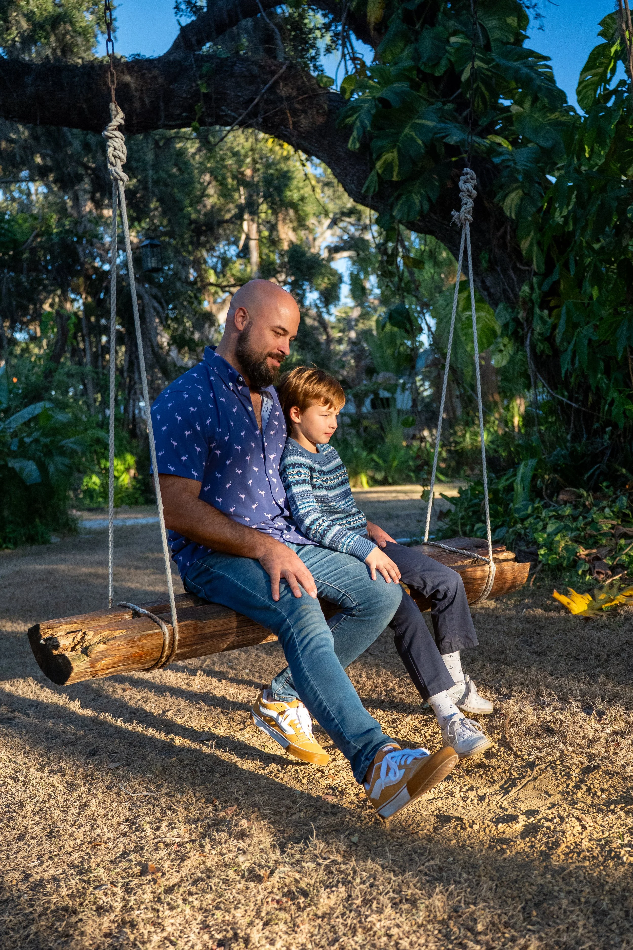 A man and a boy sitting on a wooden swing hanging from a tree in an outdoor setting during late afternoon or early evening. The man has a bald head and beard, wearing a blue shirt with a pattern, and the boy has short hair, wearing a patterned sweater. Both are looking down and smiling softly. The background features lush green foliage and a clear blue sky.