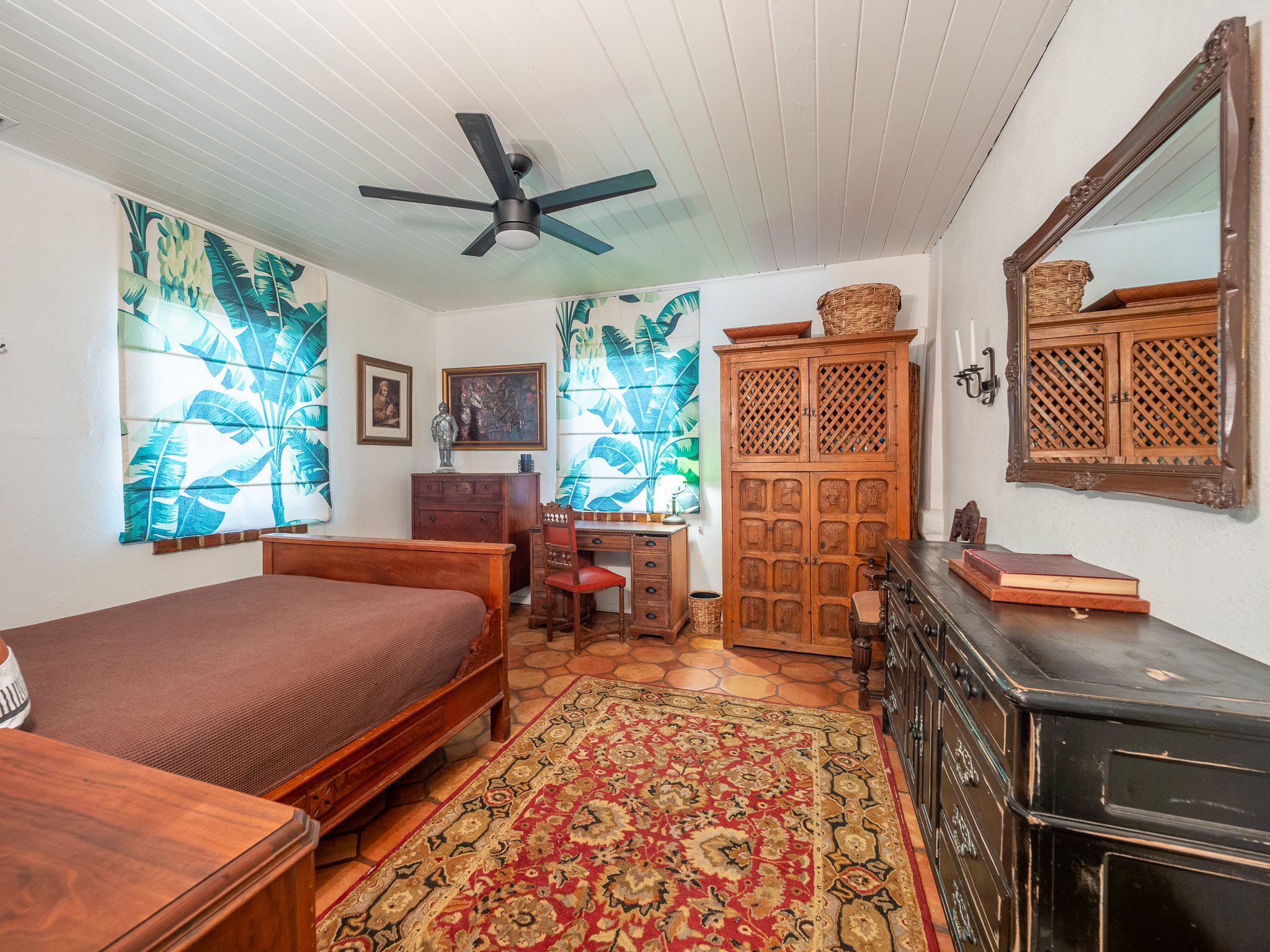 A bedroom with a white-paneled ceiling, ceiling fan, and tropical-themed curtains covering two windows. There is wooden furniture, including a small cabinet with framed pictures, a wardrobe, and a dresser. The room has a patterned rug and various dec