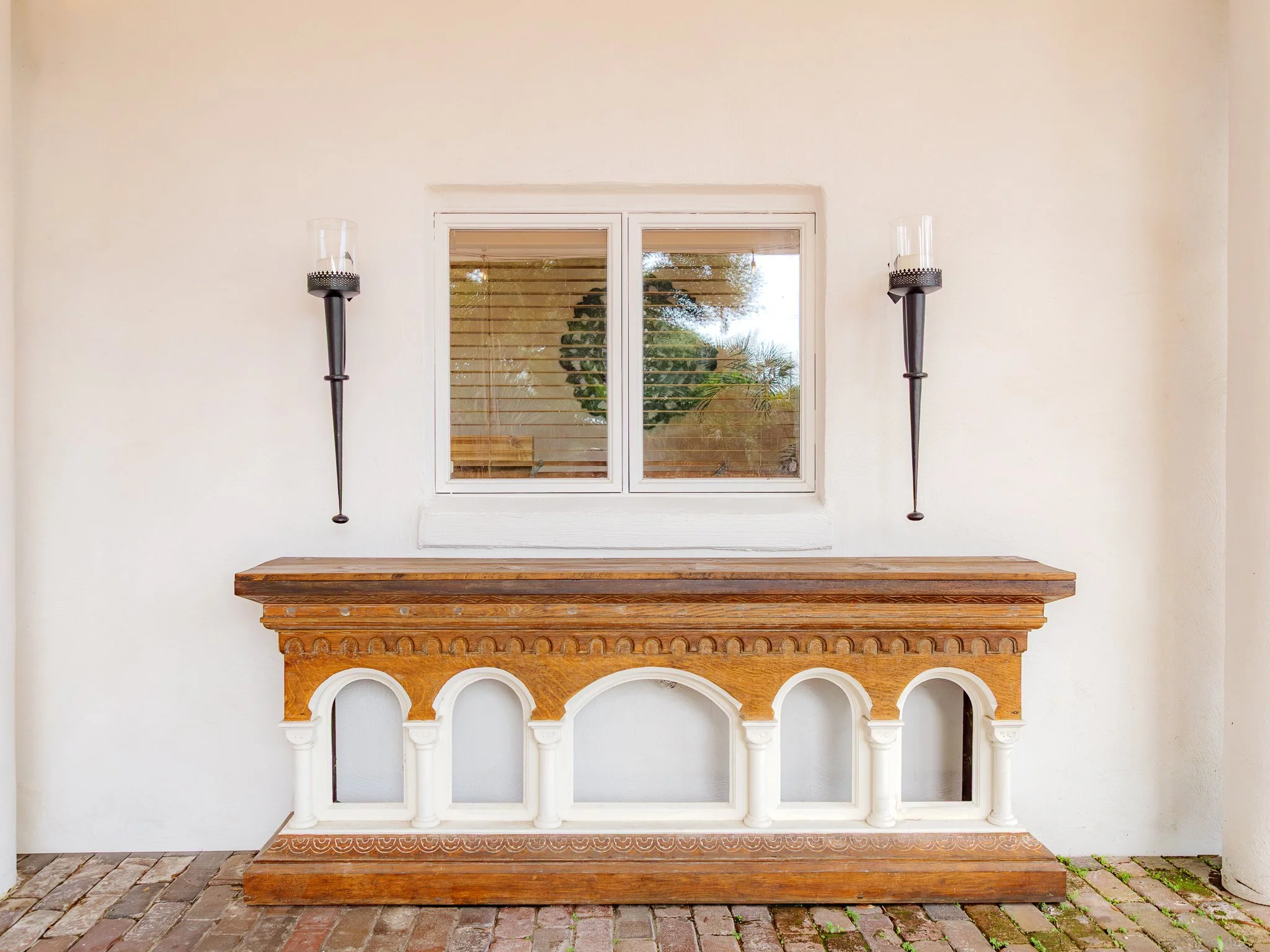 Avintage wooden mantel with ornate arches and columns, placed against a plain white wall. Two tall black sconces with glass covers are mounted on the wall on each side of a small window with wooden blinds. The window reflects trees and outdoor scener