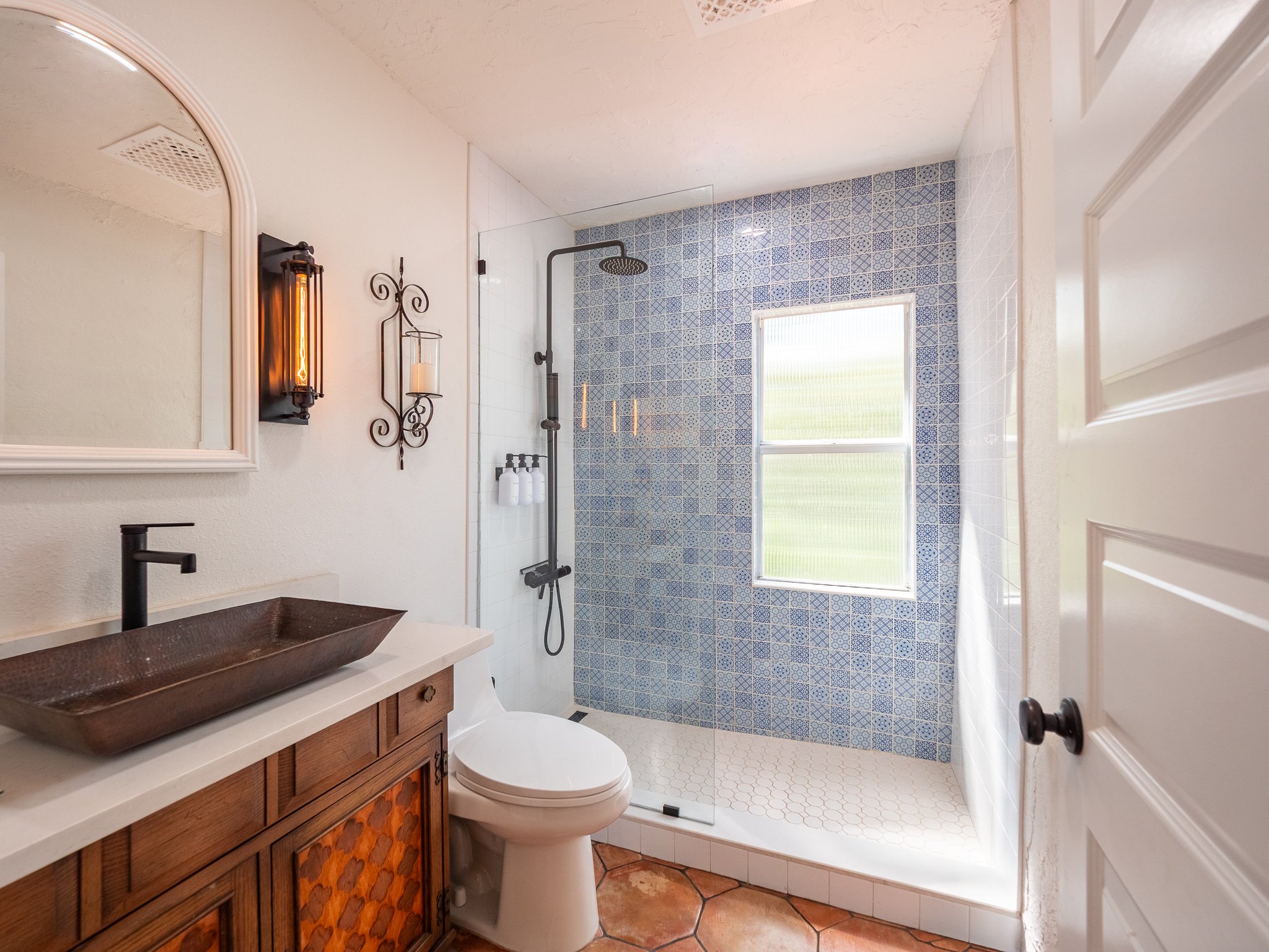 Bathroom with a shower area featuring blue patterned tiles and a rain showerhead, a window, a wooden vanity with a copper sink, a mirror, and wall sconces.