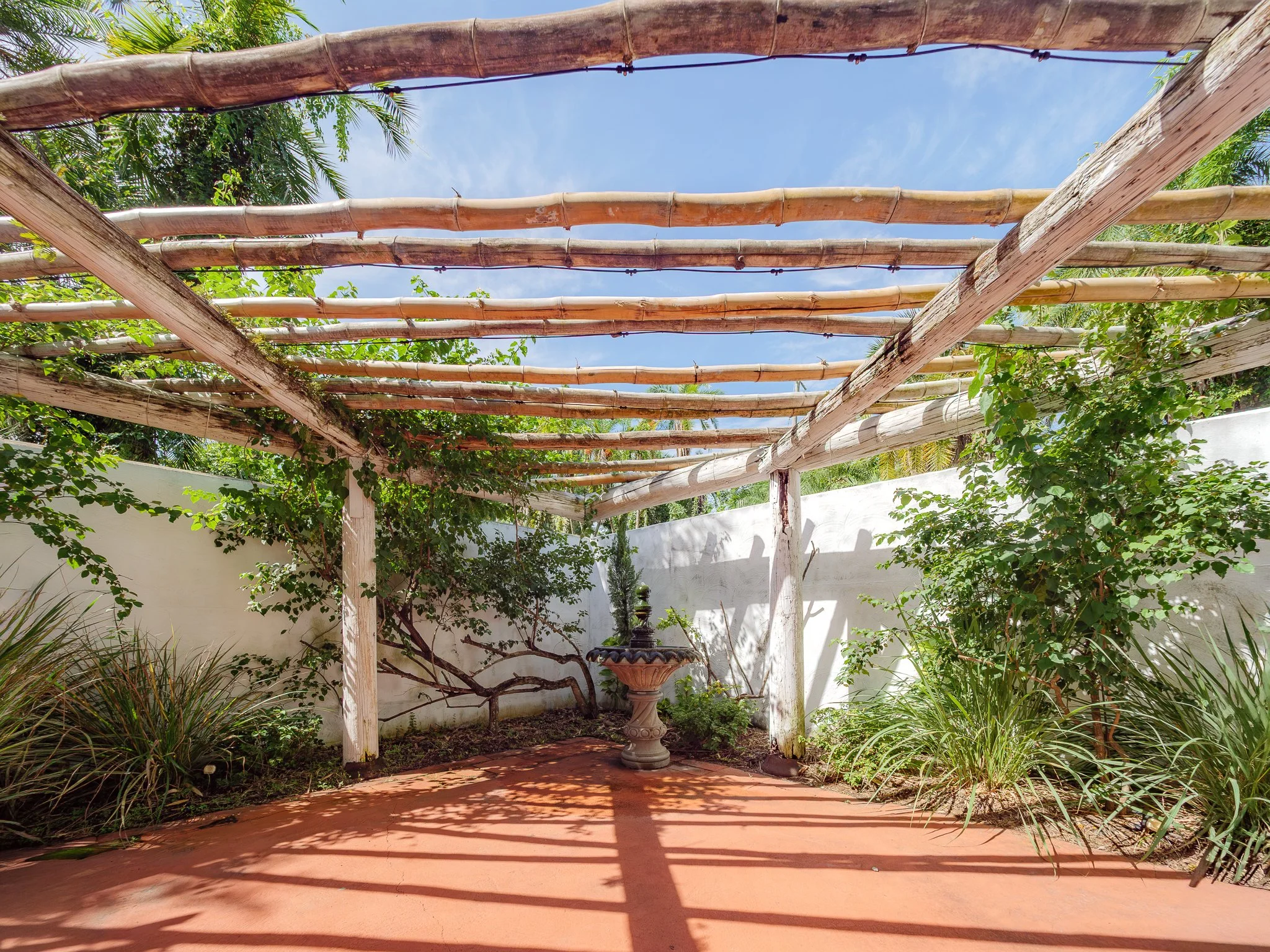 A backyard garden with a white wooden pergola overhead, surrounded by lush green plants and trees, and a sitting area with a birdbath fountain in the center. The sky is partly cloudy.