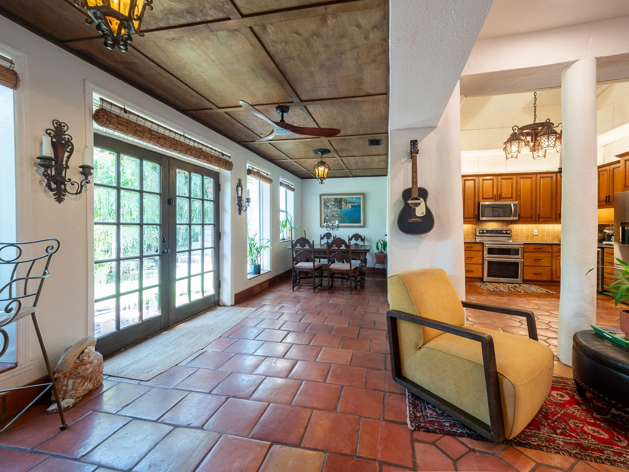Interior of a room with large windows, a wooden ceiling, hanging chandeliers, a wall-mounted ceiling fan, a guitar on the wall, and wooden furniture. The room opens to an outdoor area with greenery visible through the windows.
