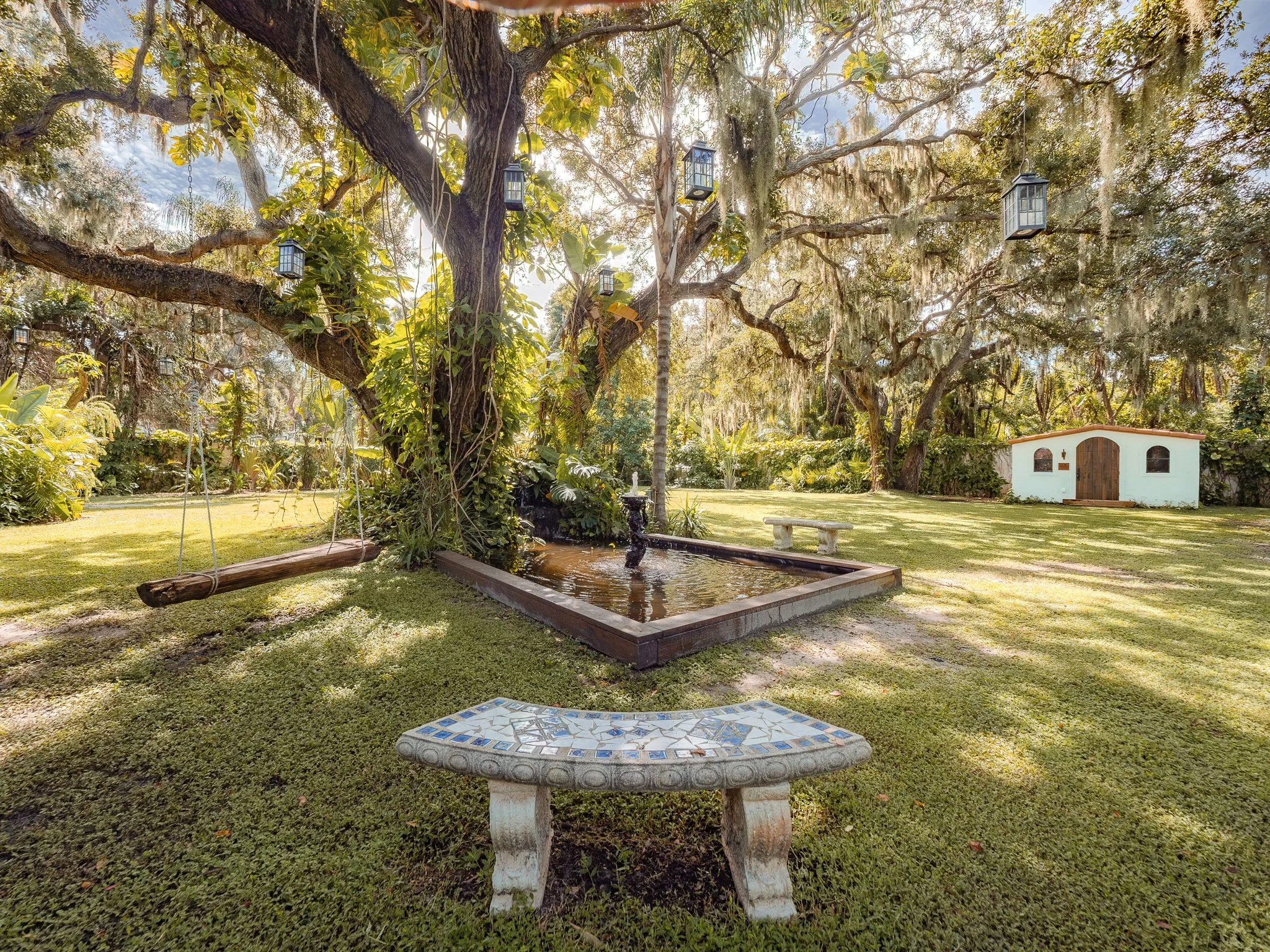 A large tree with hanging moss and lanterns hanging from its branches, a fountain inside a koi pond, a stone table, a small white casita with an arched door, and lush green plants and grass in a garden.