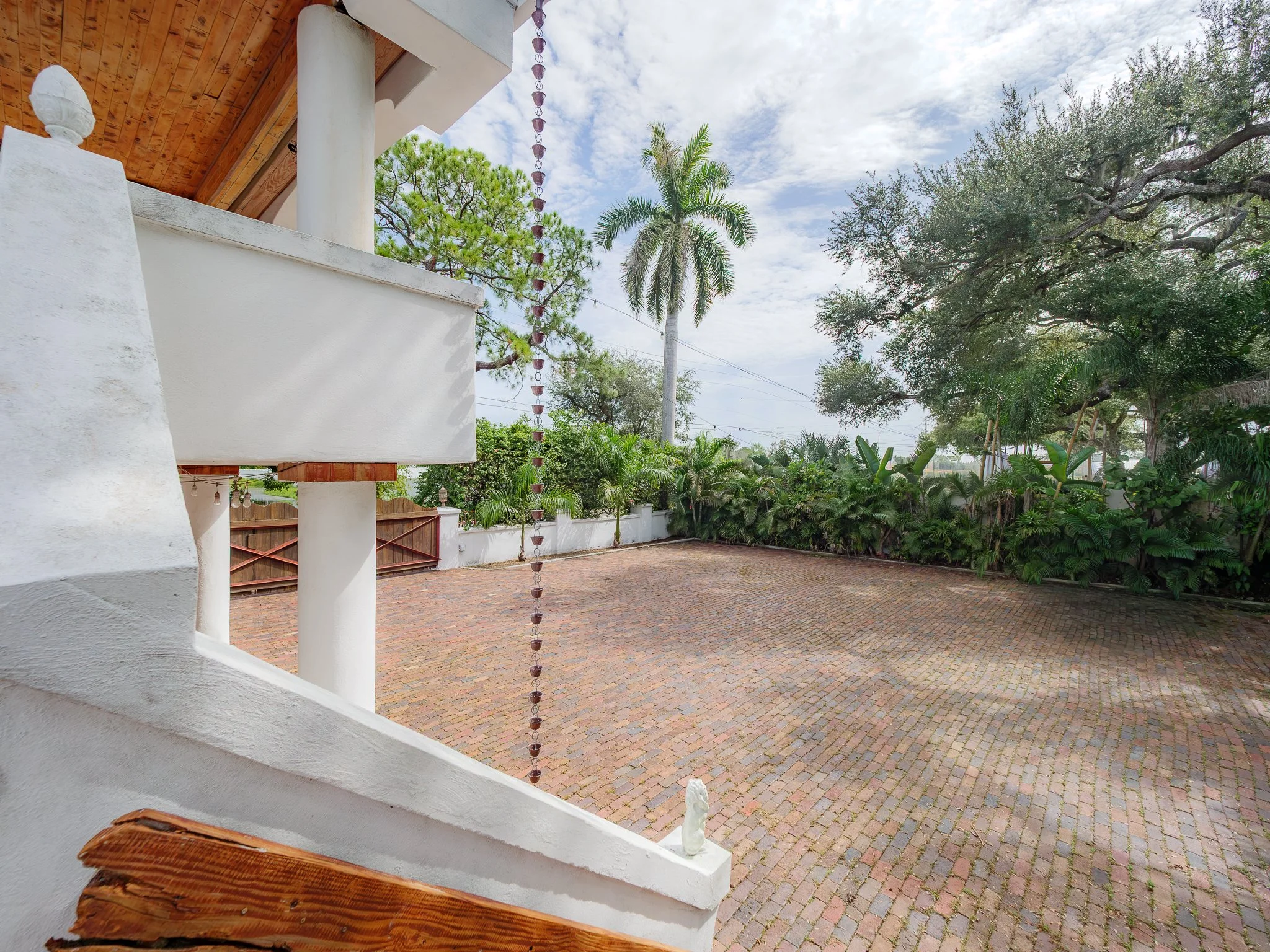 View from a porch with a white railing, showing a paved courtyard, various trees including a palm and pine, and a cloudy sky.