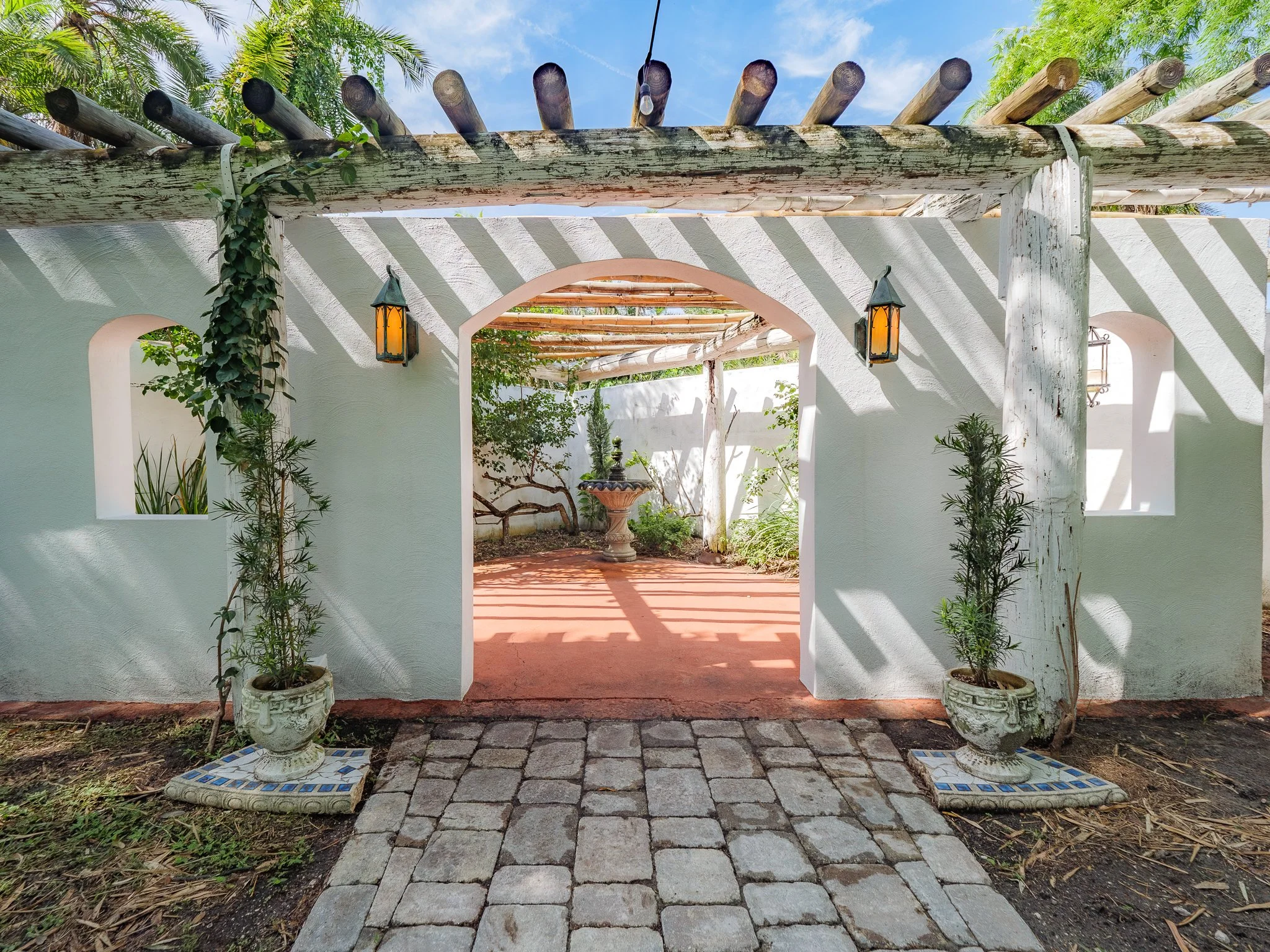 A patio scene viewed through an arched opening framed by white stucco walls. The patio features two potted plants on either side of the arch and a stone pathway leading to a fountain with greenery in the background. The roof has wooden beams casting 