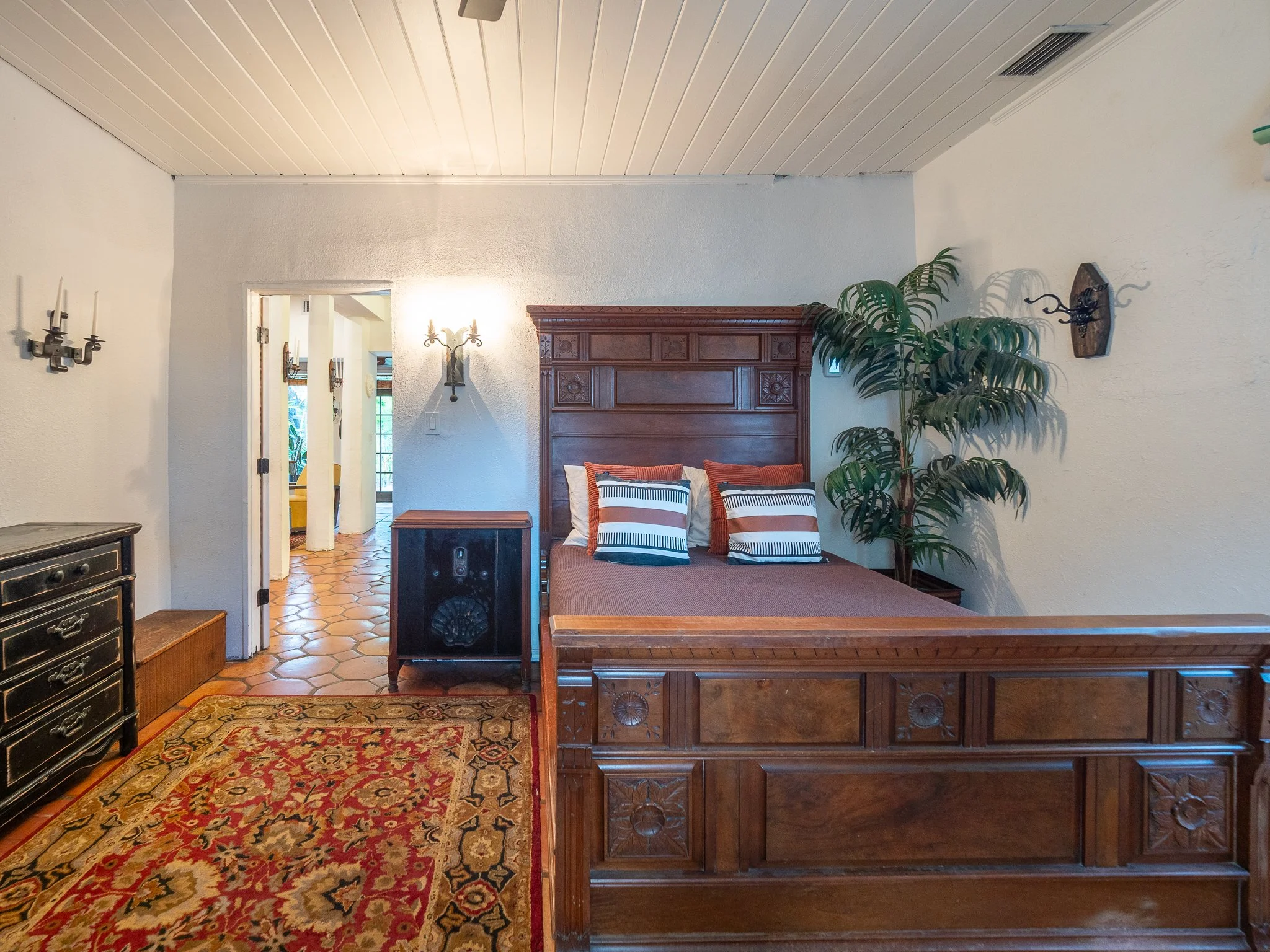 Interior of a room with beige walls and a white wooden ceiling. There is a wooden headboard with carved details, striped cushions, a large potted plant, a vintage cabinet, a decorative rug, and wall sconces with lit candles.