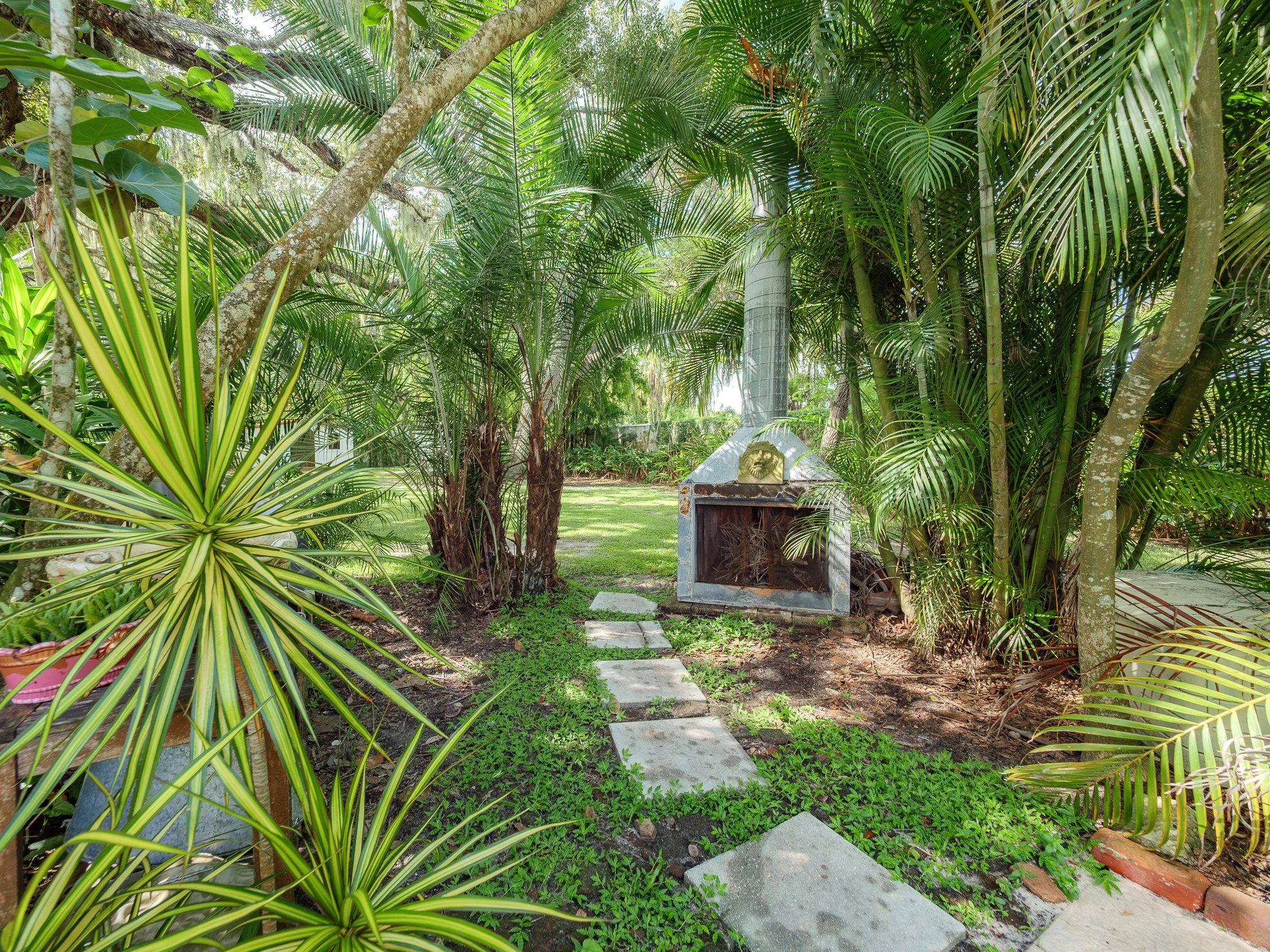 A lush tropical garden with green plants, trees, and a stone pathway leading to a small shrine with a lion sculpture on top, surrounded by dense foliage.