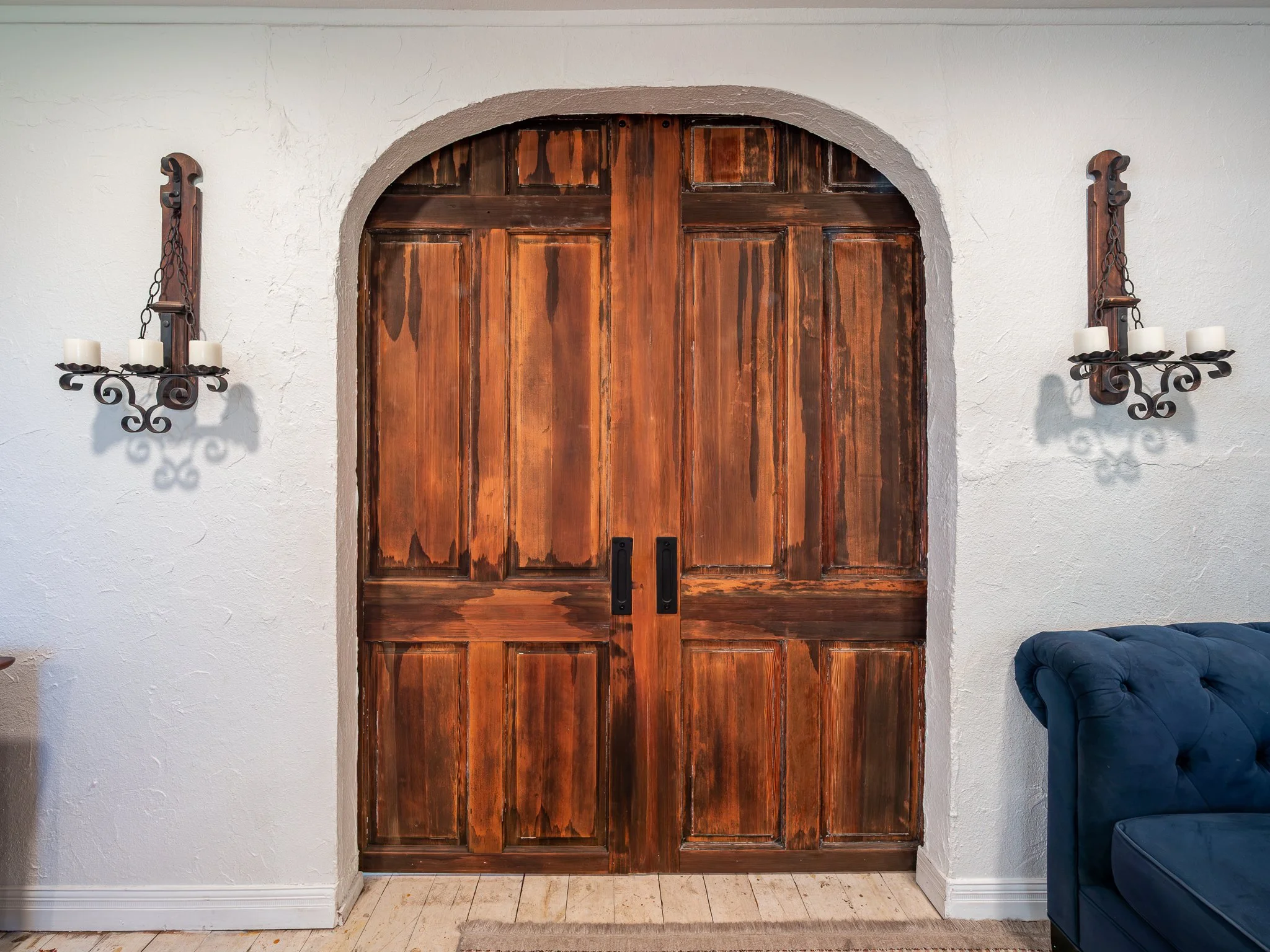 Large wooden double doors with black handles and decorative black wall sconces with candles on either side, set within a white stucco wall.