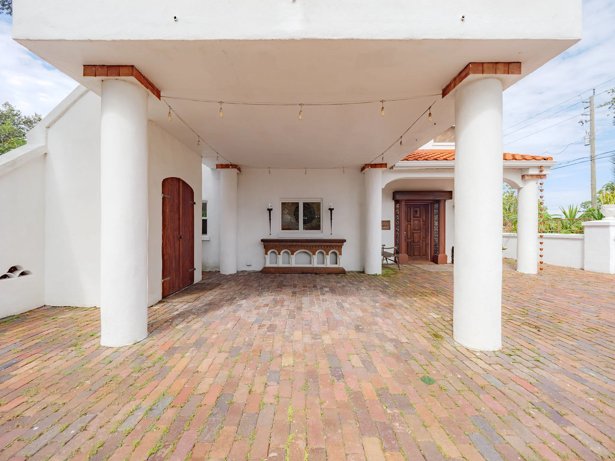 Front balcony of a house with white stucco walls, large white pillars, a brick walkway, wooden doors, and string lights hanging overhead.