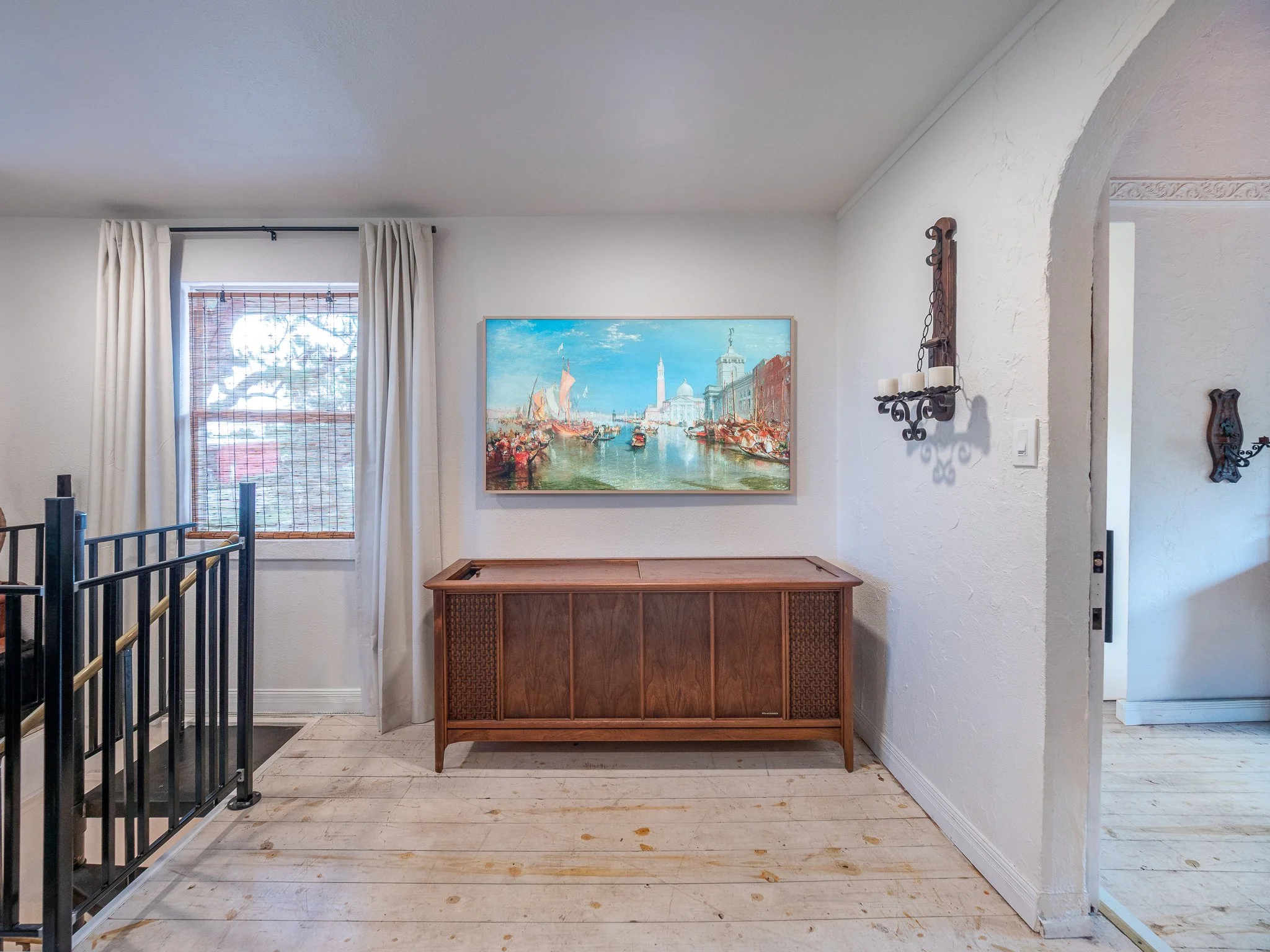 Interior of a room with white walls and light-colored wooden floor, featuring a painting of a harbor scene and a wooden sideboard.