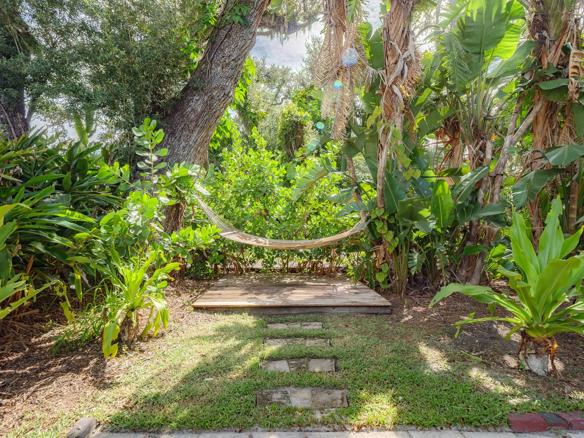 A hammock hanging between two trees in a lush jungle garden with grass and a stone pathway in the foreground.