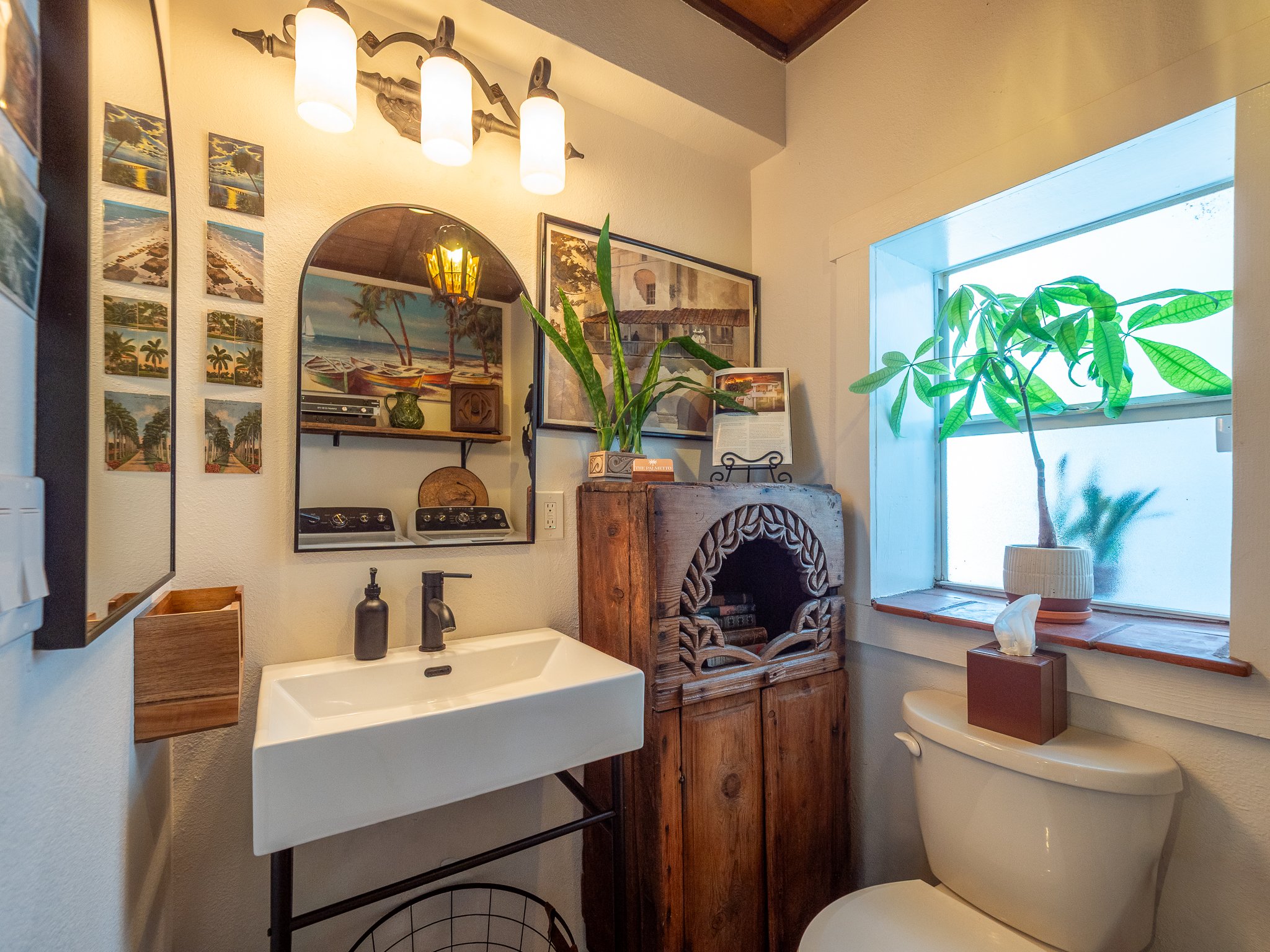 A small bathroom with a white wall-mounted sink, black faucet, and decorative wooden container on the side. Above the sink, a mirror reflects some tropical artwork and a small shelf with a green vase. To the right, there is a wooden cabinet with carv