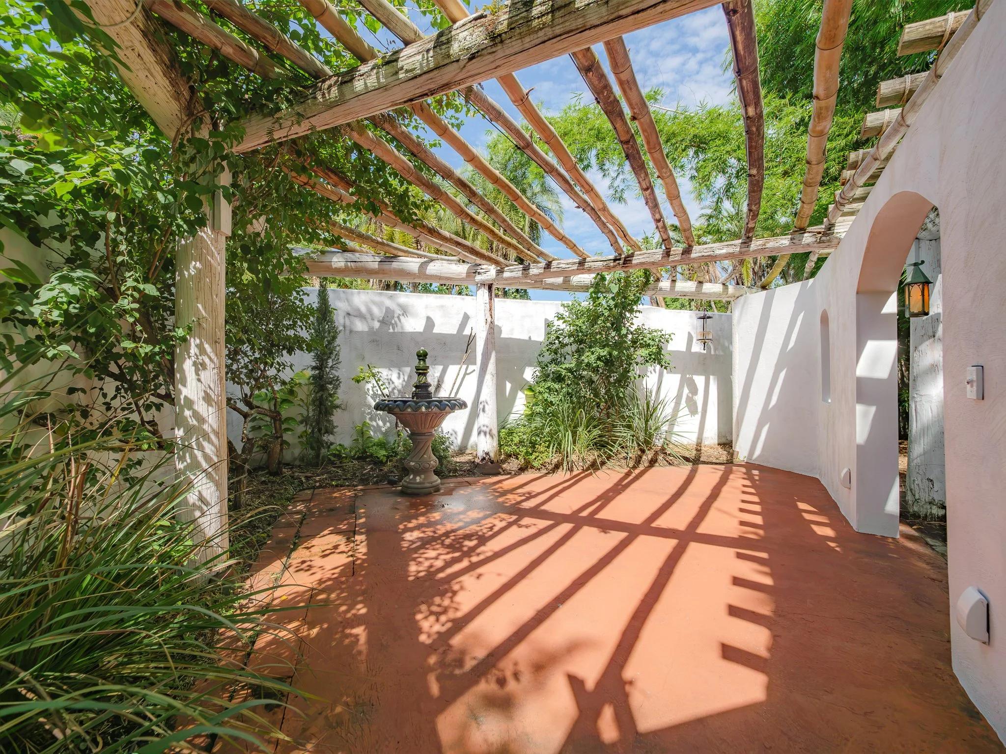 An outdoor patio area with terracotta color flooring, white stucco walls, green plants, a decorative fountain, and wooden pergola beams with climbing vines casting shadows.