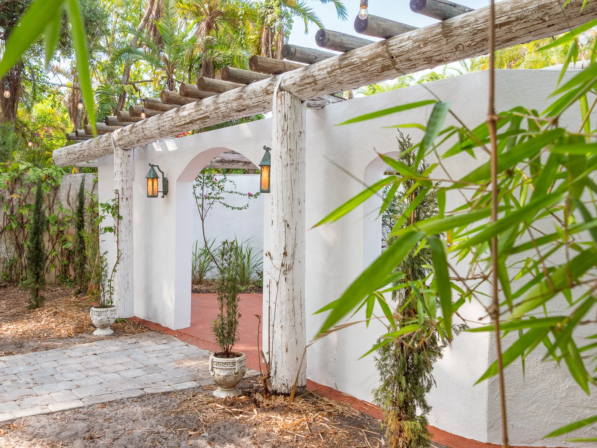 A white stucco archway with two decorative lanterns hanging on either side, surrounded by lush green plants and trees with a bamboo plant visible in the foreground and a tree with hanging light bulbs in the background.