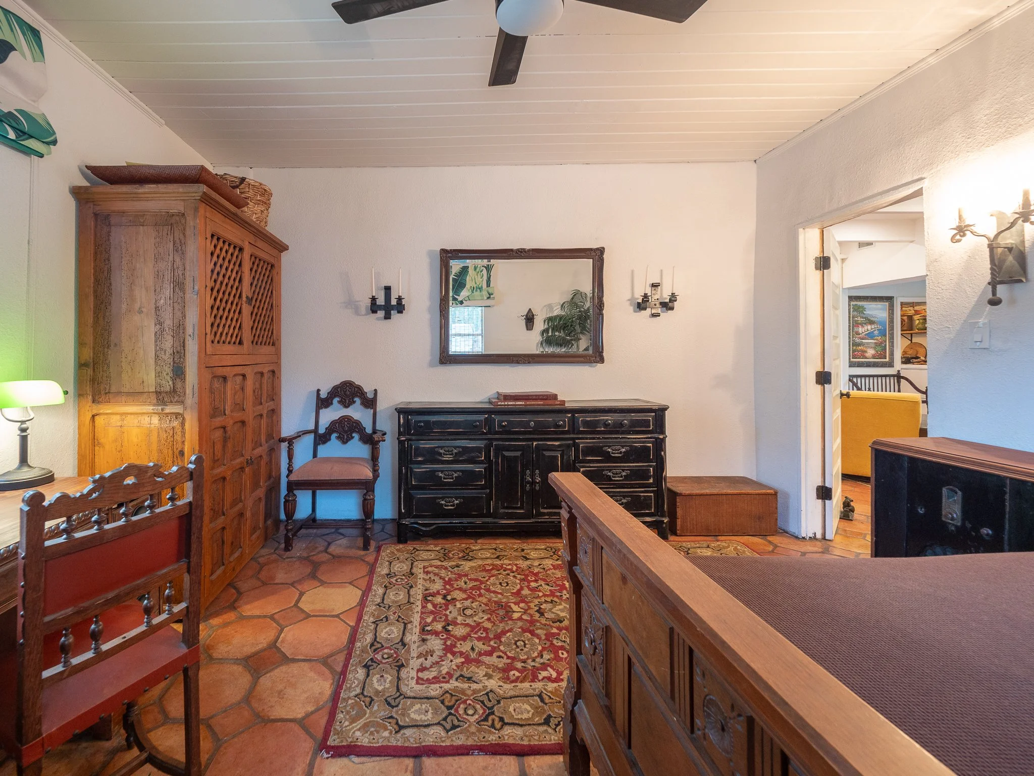 The image shows a cozy room with wooden furniture, a mirror on the wall, a black dresser, a patterned rug, and a doorway leading to another room decorated with paintings, a yellow sofa, and a railing.