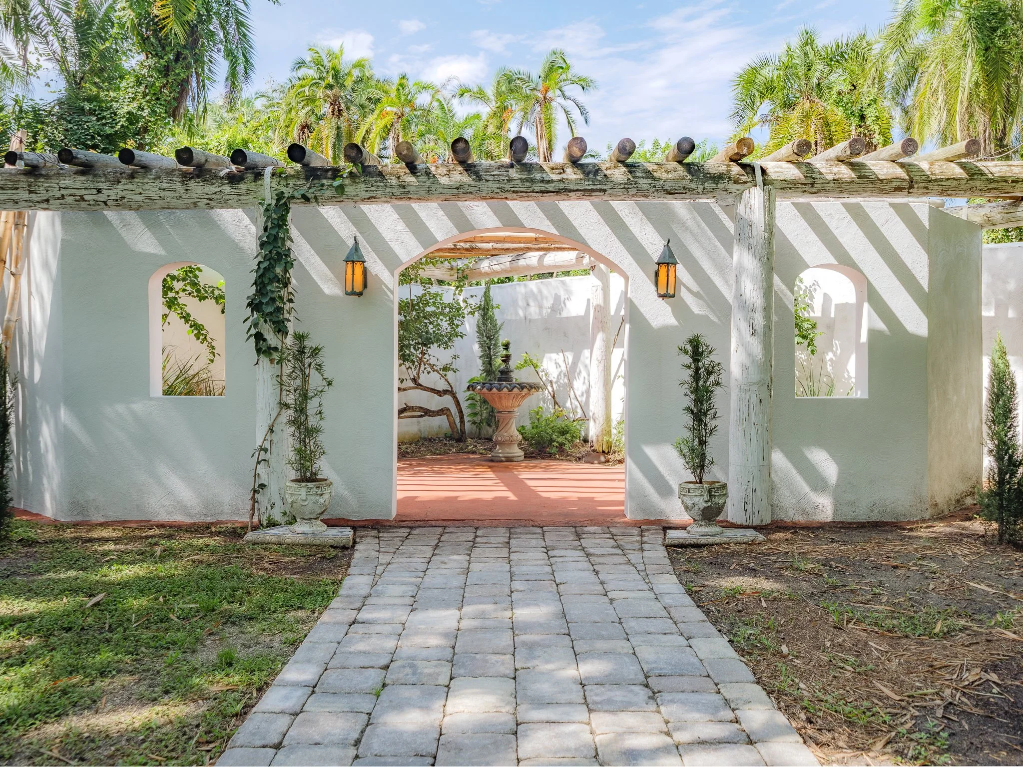 A white stucco wall with an arched opening, flanked by vintage lantern-style lights, and a stone pathway leading into a lush garden with tropical trees and plants.