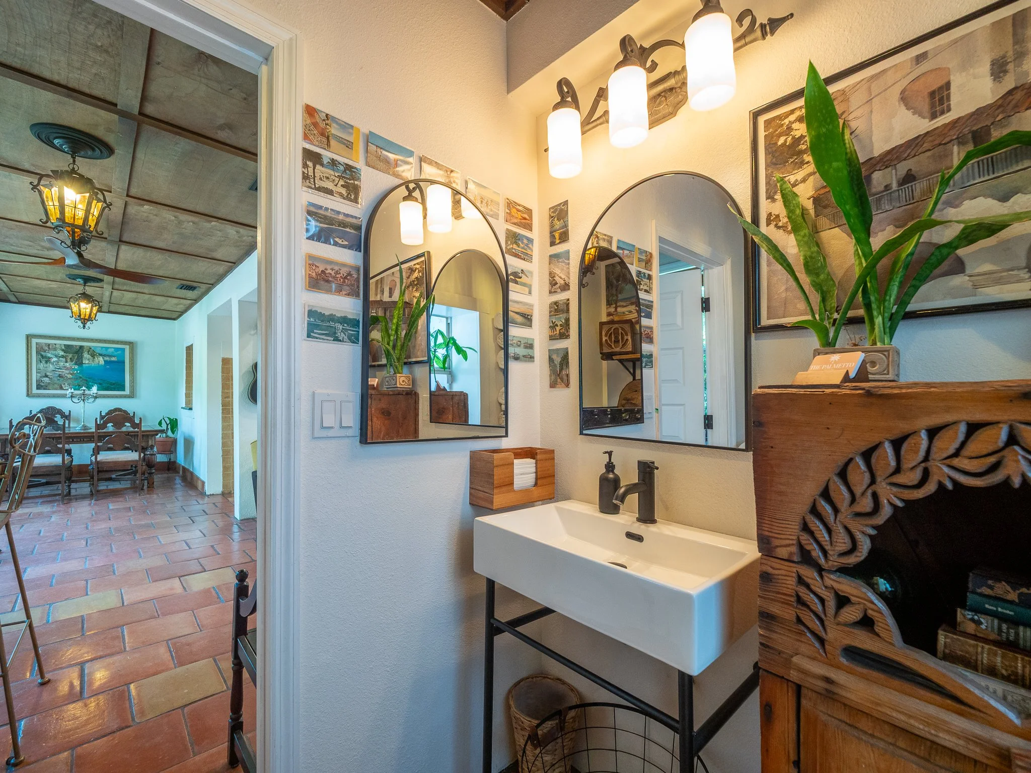 Interior view of a cozy bathroom with a white sink and black faucet, decorative mirrors, and potted plants. The hallway has a gallery wall with pictures and a large framed artwork, with warm lighting from ceiling fixtures. The adjoining room has a wo