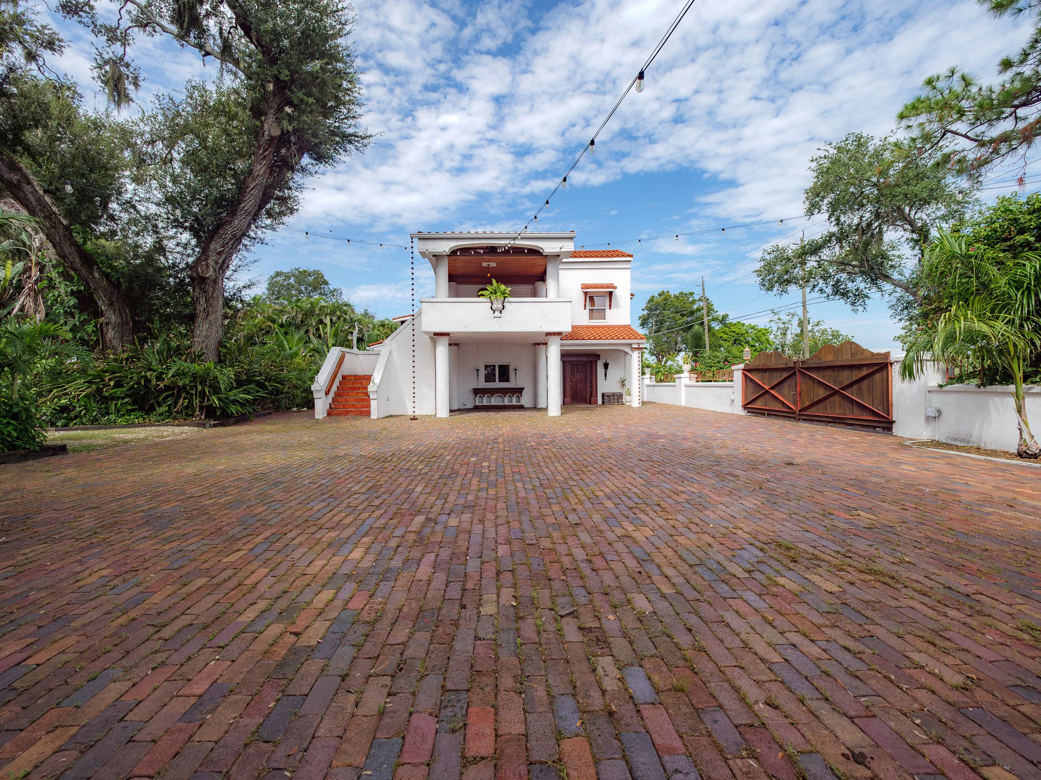 A white two-story house with a Spanish-tiled roof, an outdoor balcony with potted plants, a wooden front door, and a large brick courtyard surrounded by lush greenery and tall trees, under a partly cloudy sky.