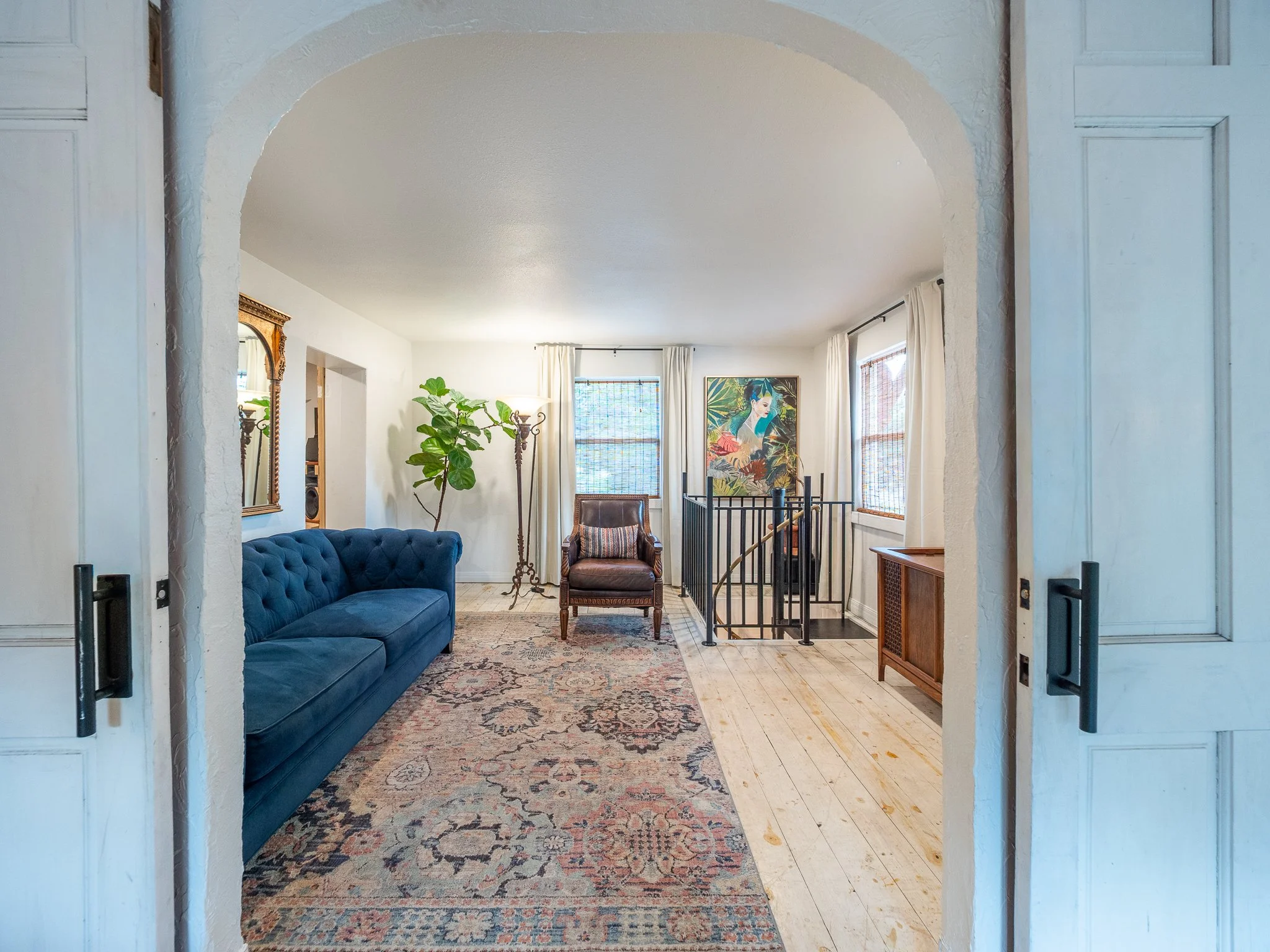 Living room with a blue couch, a patterned rug, a potted plant, a cushioned chair, a framed artwork of a woman, and windows with white curtains, viewed through an arched doorway.