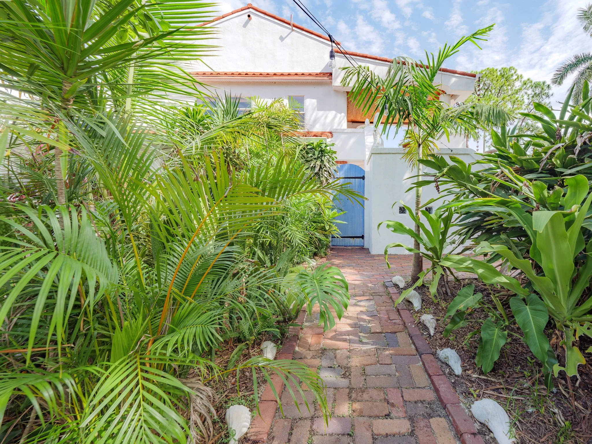 A white house with a red-tile roof and clay tiles on the side, surrounded by tropical plants and trees. A blue door is visible and a brick pathway leads to it. String lights are hanging above, and the sky is partly cloudy with sunlight.