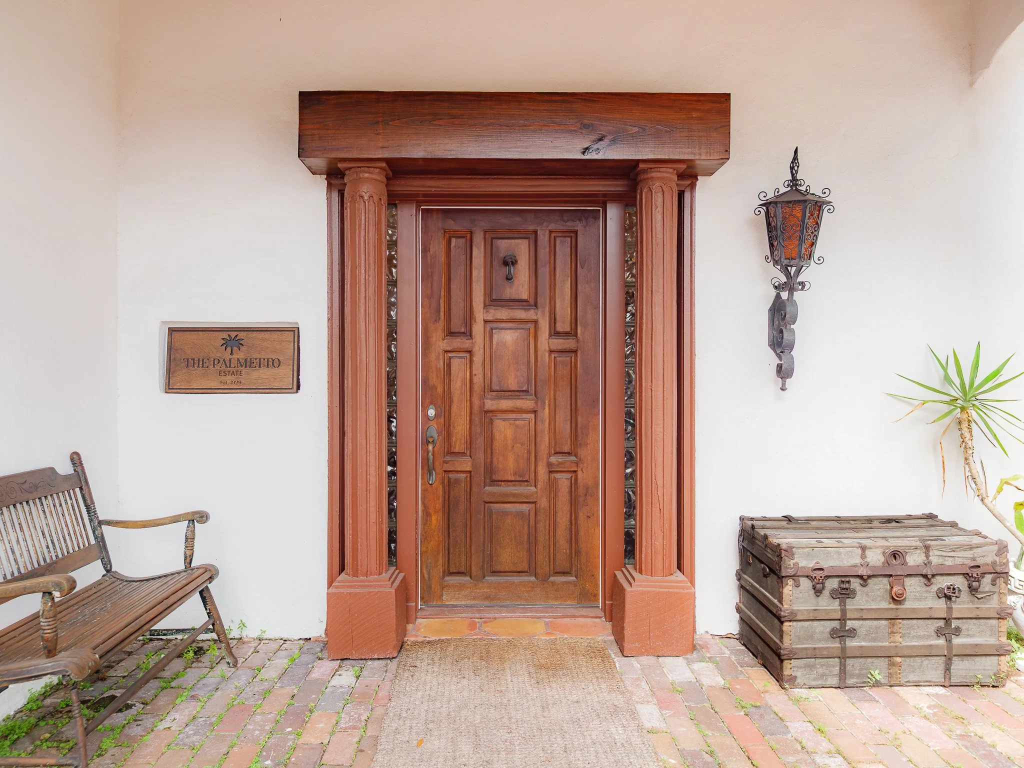 The front door of a house with a wooden finish, flanked by columns, with a black lantern-style light fixture on the right side. There is a small sign on the left that reads 'The Palmetto Estate Est. 1929'. A wooden bench is visible on the left side, 