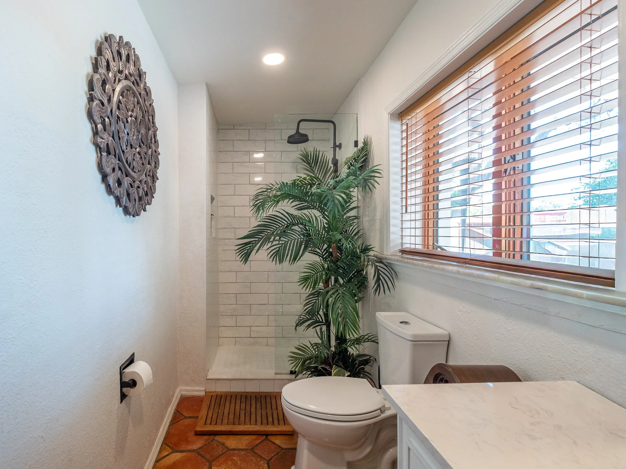 A small bathroom with white walls and terracotta tile flooring. There is a white toilet and a wooden bath mat in front of it. A large window with wooden blinds lets in natural light. An indoor plant and a decorative wooden wall art piece are on the w