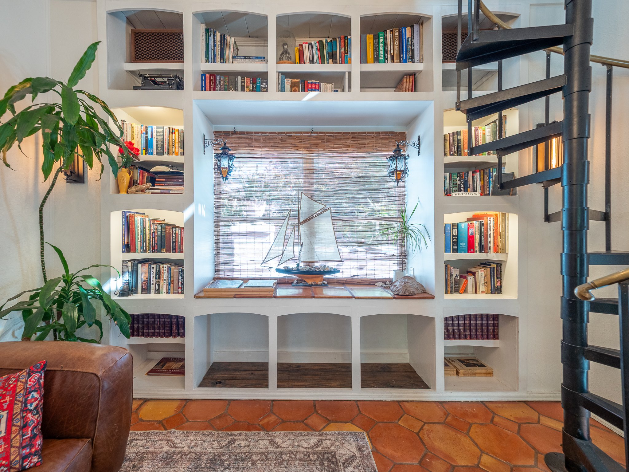 A white built-in bookshelf with various books, decorative items, and a vintage typewriter on one shelf, located in a living room with a large window, bamboo shades, hanging lanterns, a potted plant, and a staircase to the right.