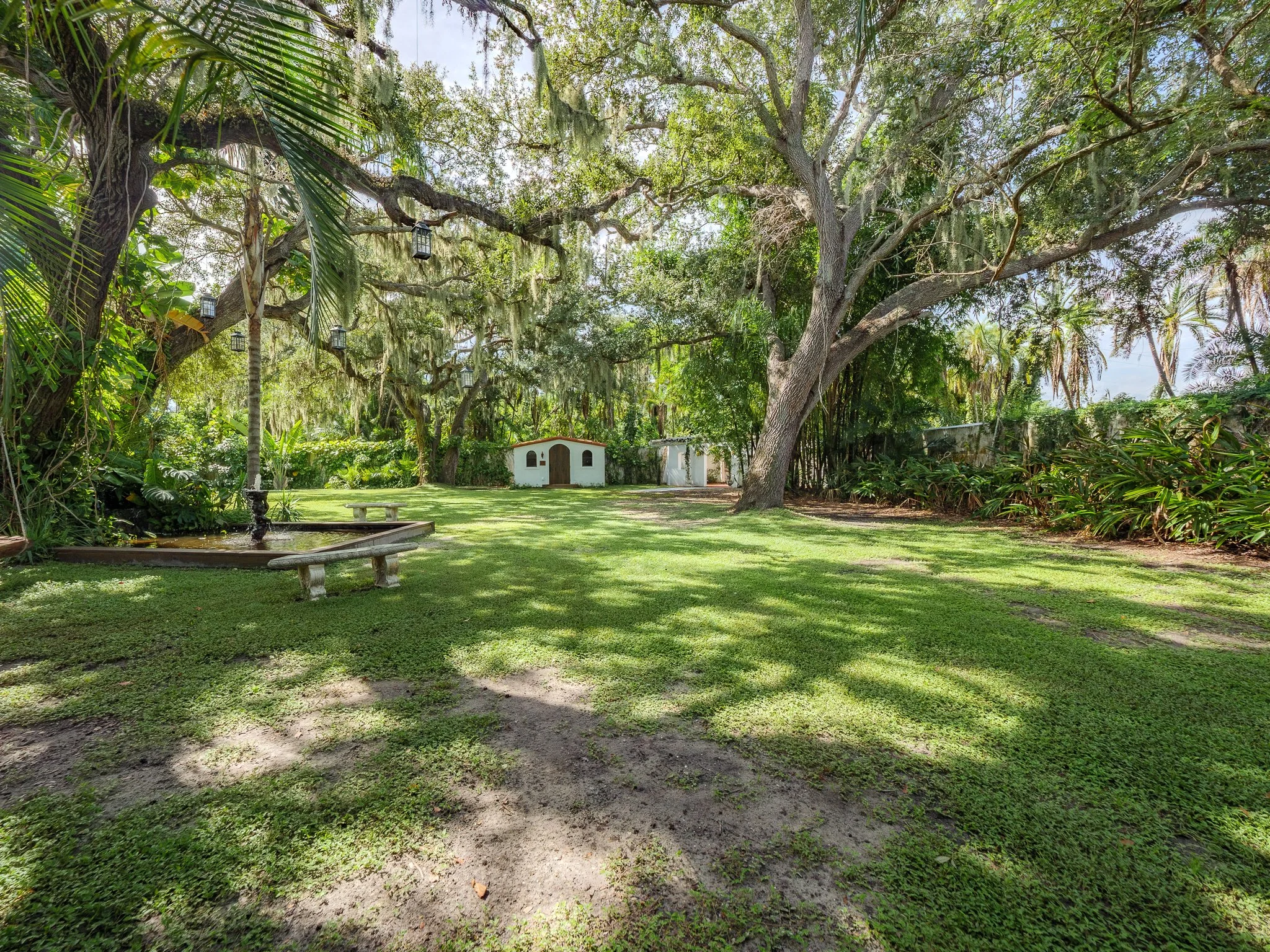 A lush green garden with large trees, a small white building, a bench, and a fountain, under a partly cloudy sky.