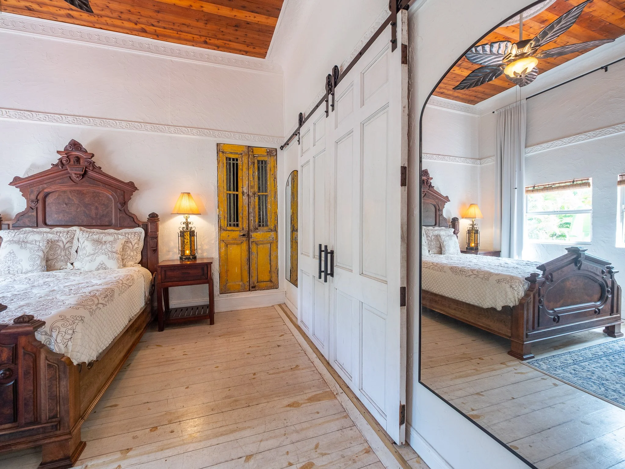 A rustic bedroom with a large wooden bed, ornate headboard, white bedding with a pattern, two bedside lamps, a yellow distressed wooden door, and a sliding barn door. The room has wooden floors, a wooden ceiling, and a window with a curtain.