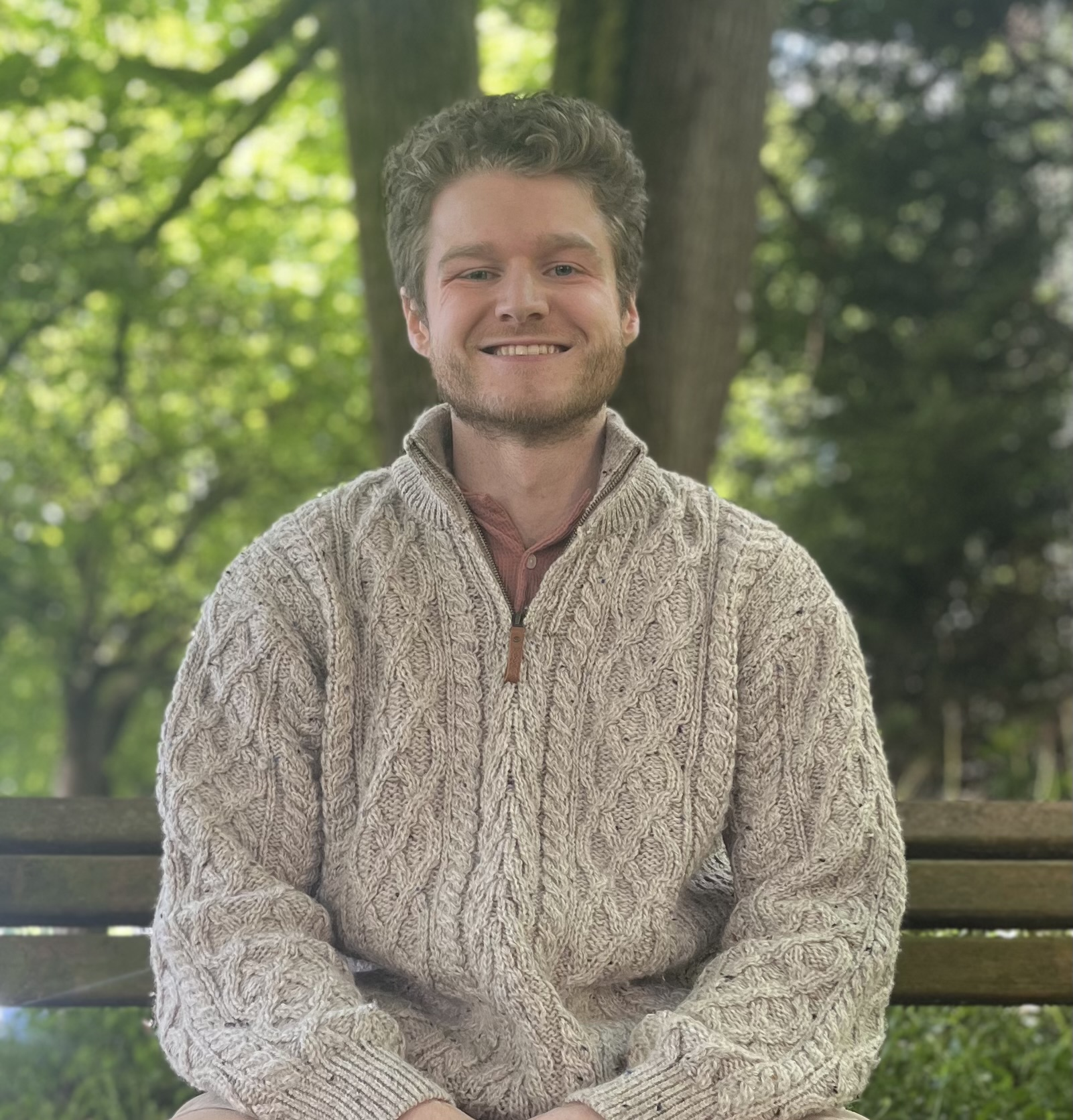 A young man with curly hair and a beard sitting on a bench outdoors, wearing a beige cable-knit sweater and smiling, with green trees in the background.