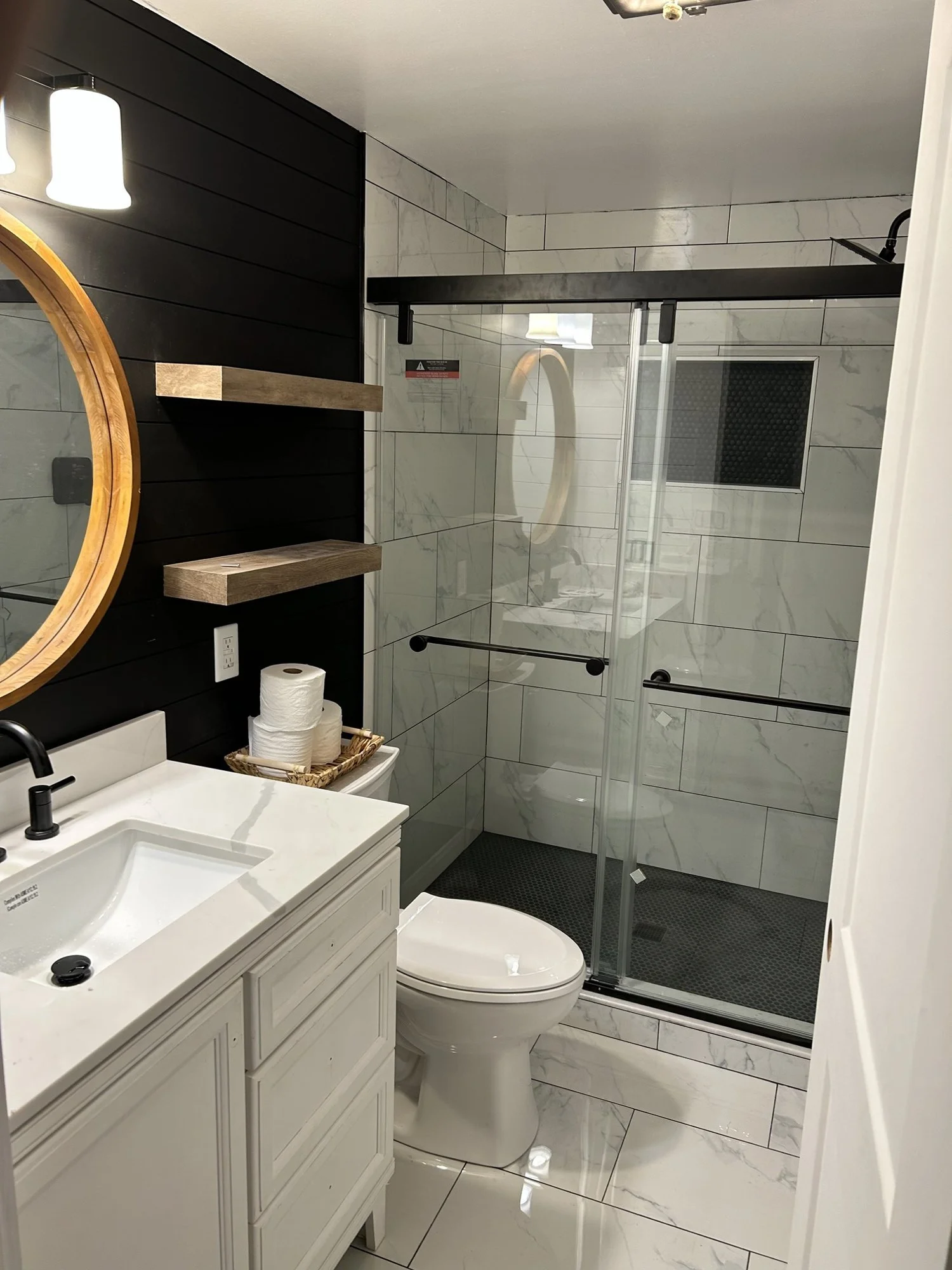 Bathroom remodel: walk-in tiled shower with sliding glass door, shiplap accent wall, floating shelves and round mirror.