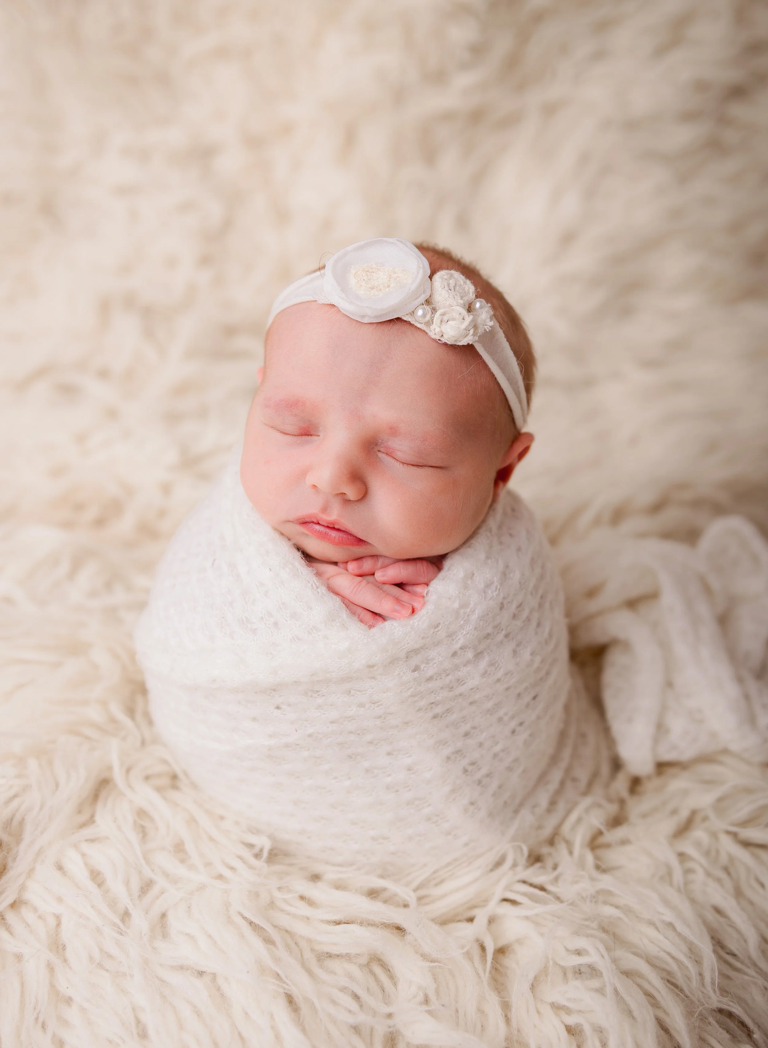 A sleeping baby wrapped in a white blanket, wearing a white headband with a decorative flower and pearls, lying on a cream-colored furry blanket.