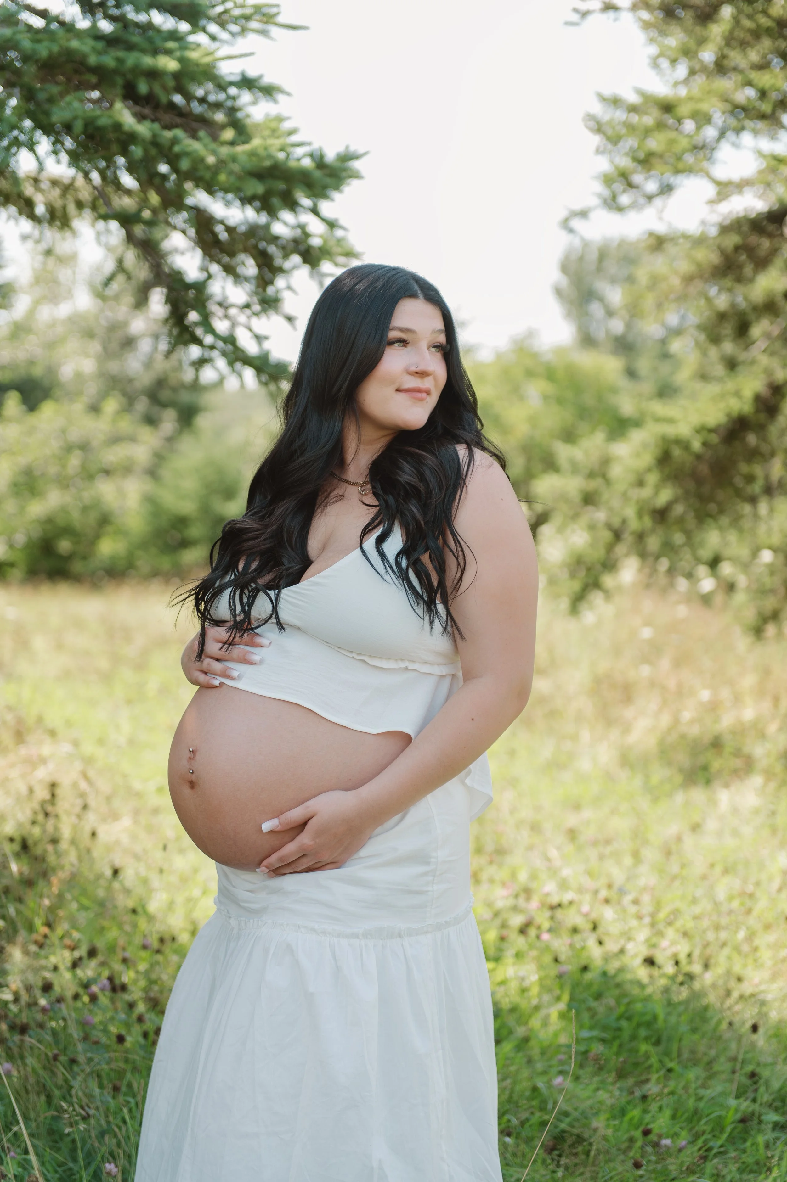 A pregnant woman with long black hair and a nose piercing is standing outdoors in a green, wooded area. She is wearing a white sleeveless top and a long white skirt, and she is holding her pregnant belly with one hand while the other rests gently on her hip.