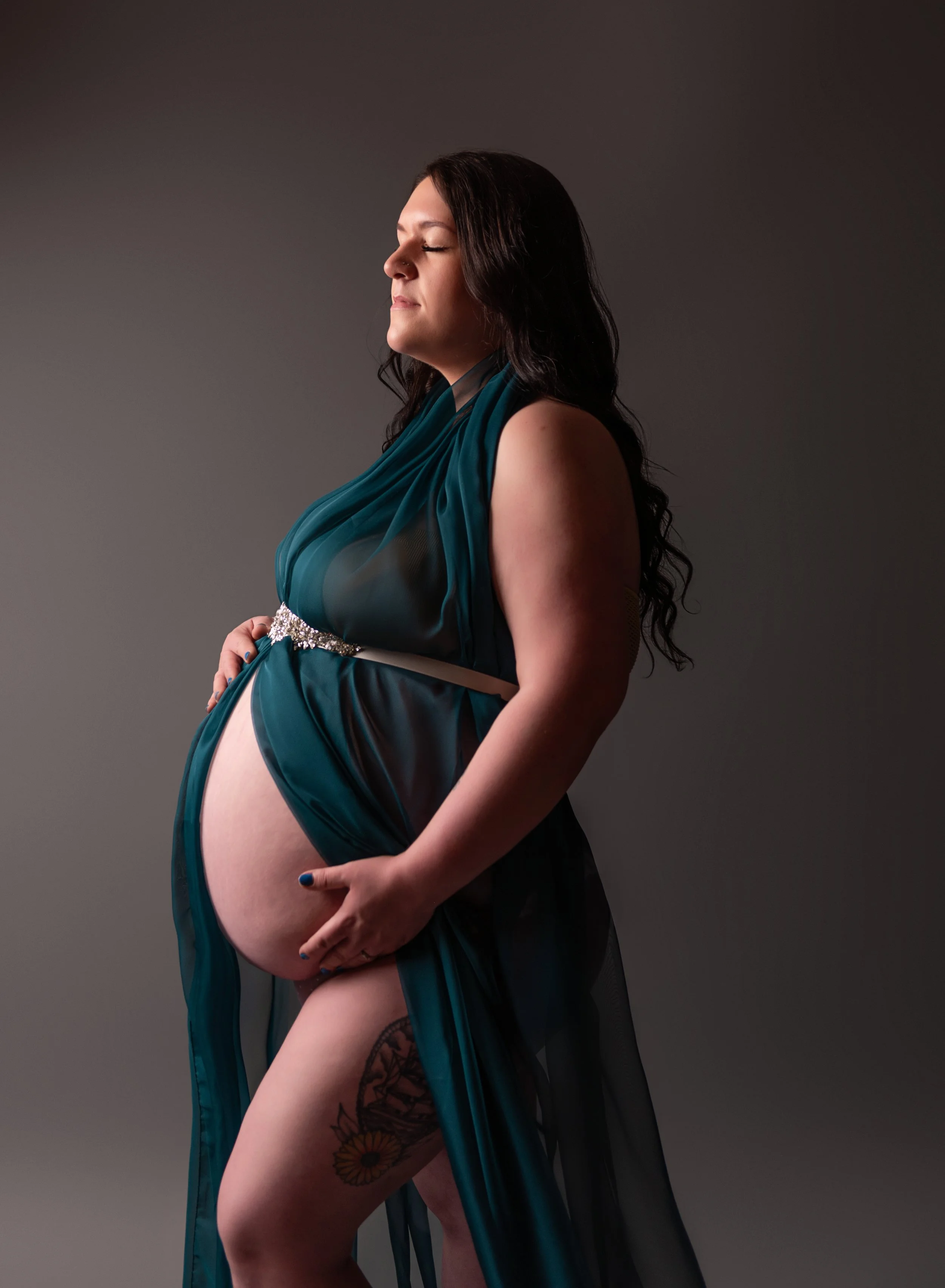 A pregnant woman standing sideways with her eyes closed, wearing a teal and black dress, with her hands resting on her belly against a dark grey background.