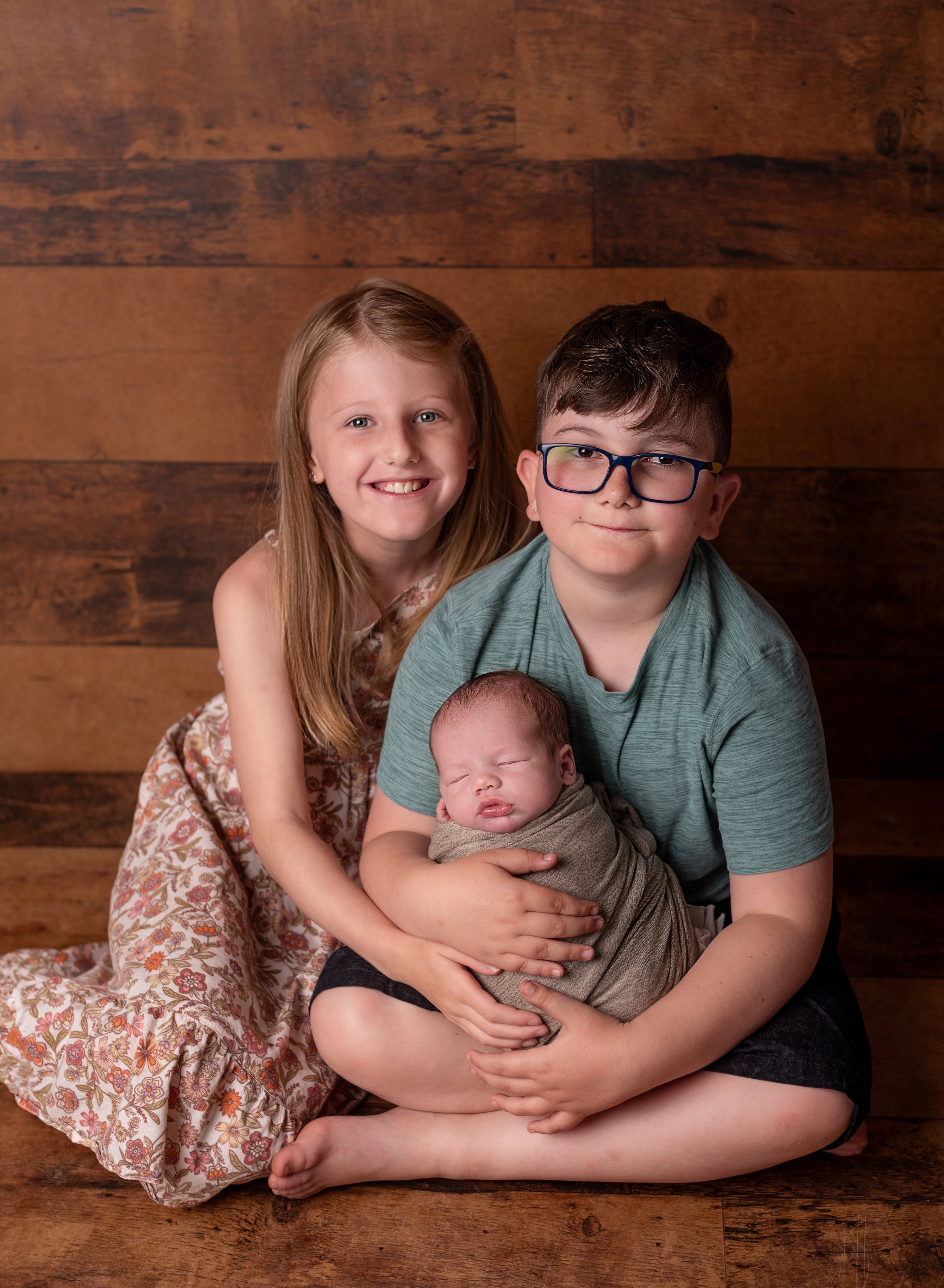 a girl and a boy sit on a wood backdrop holding their newborn sibling for a sibling shot in studio