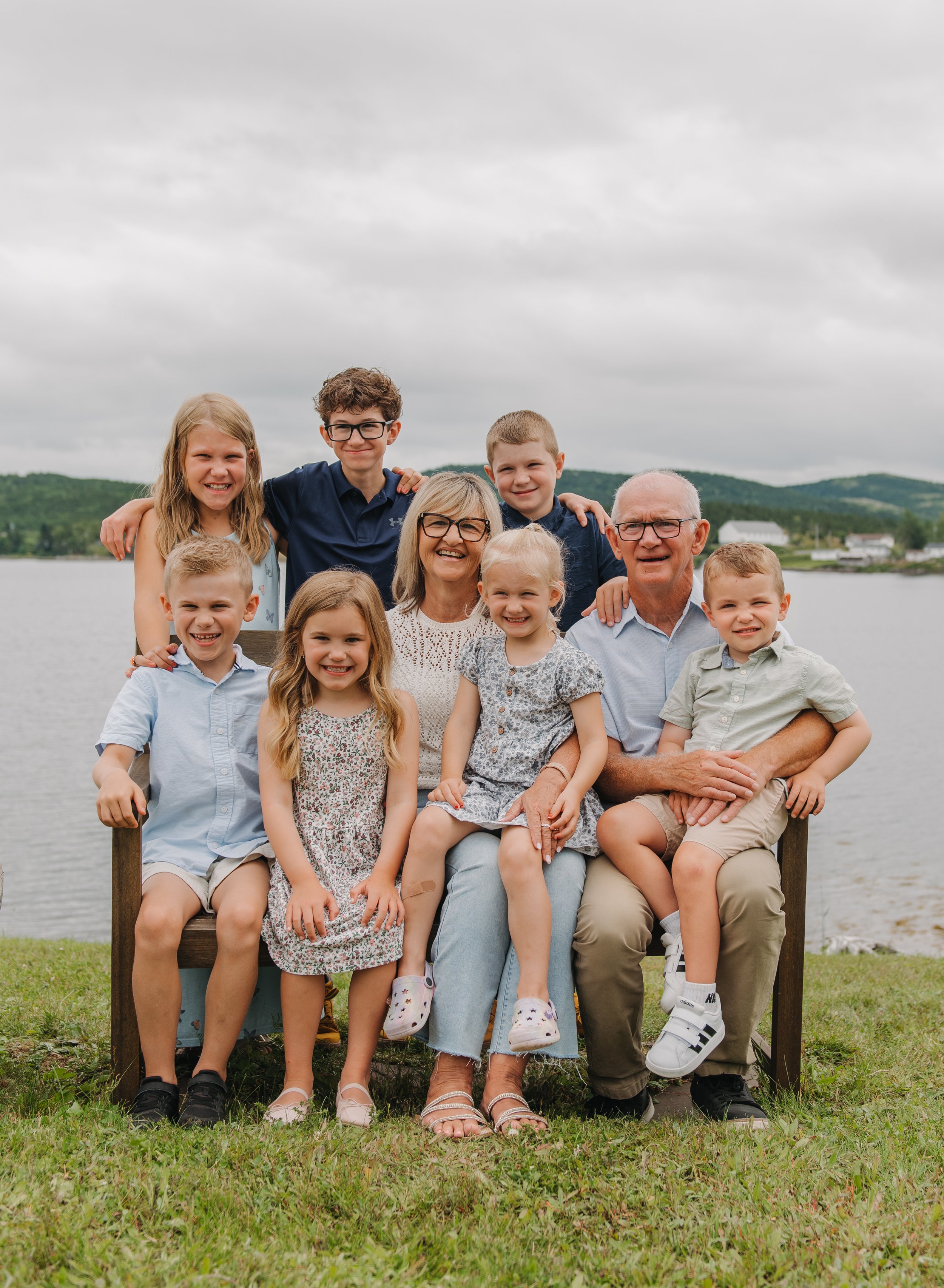 A multigenerational family of twelve people, including children, parents, and grandparents, posing outdoors at a lake with green hills in the background under cloudy skies.