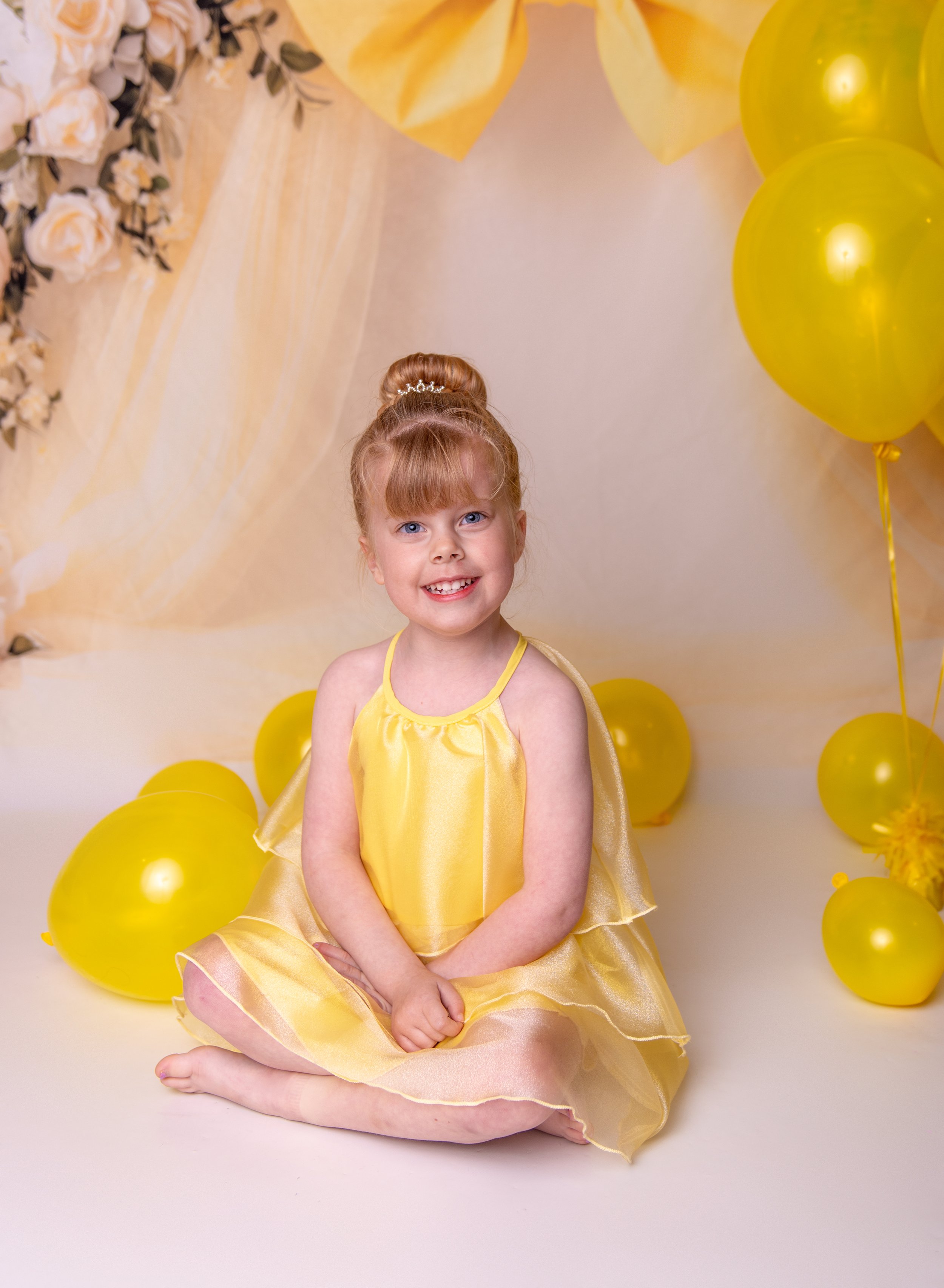 Young girl sitting on the floor, wearing a yellow dress, surrounded by yellow balloons, with a background of yellow flowers and drapery.