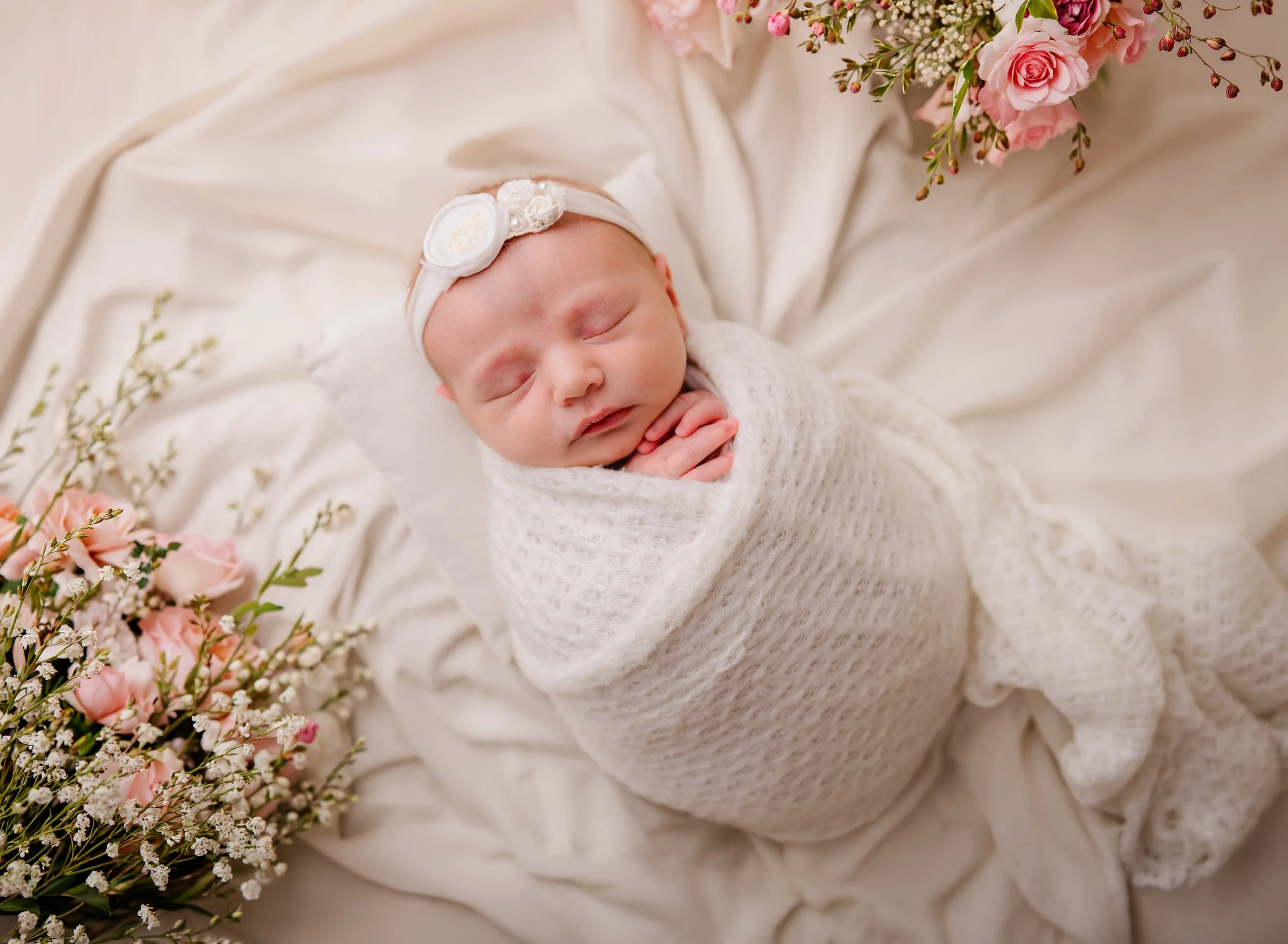 A newborn baby sleeping peacefully, wrapped in a soft white blanket, wearing a delicate headband, surrounded by pink and white flowers and greenery.