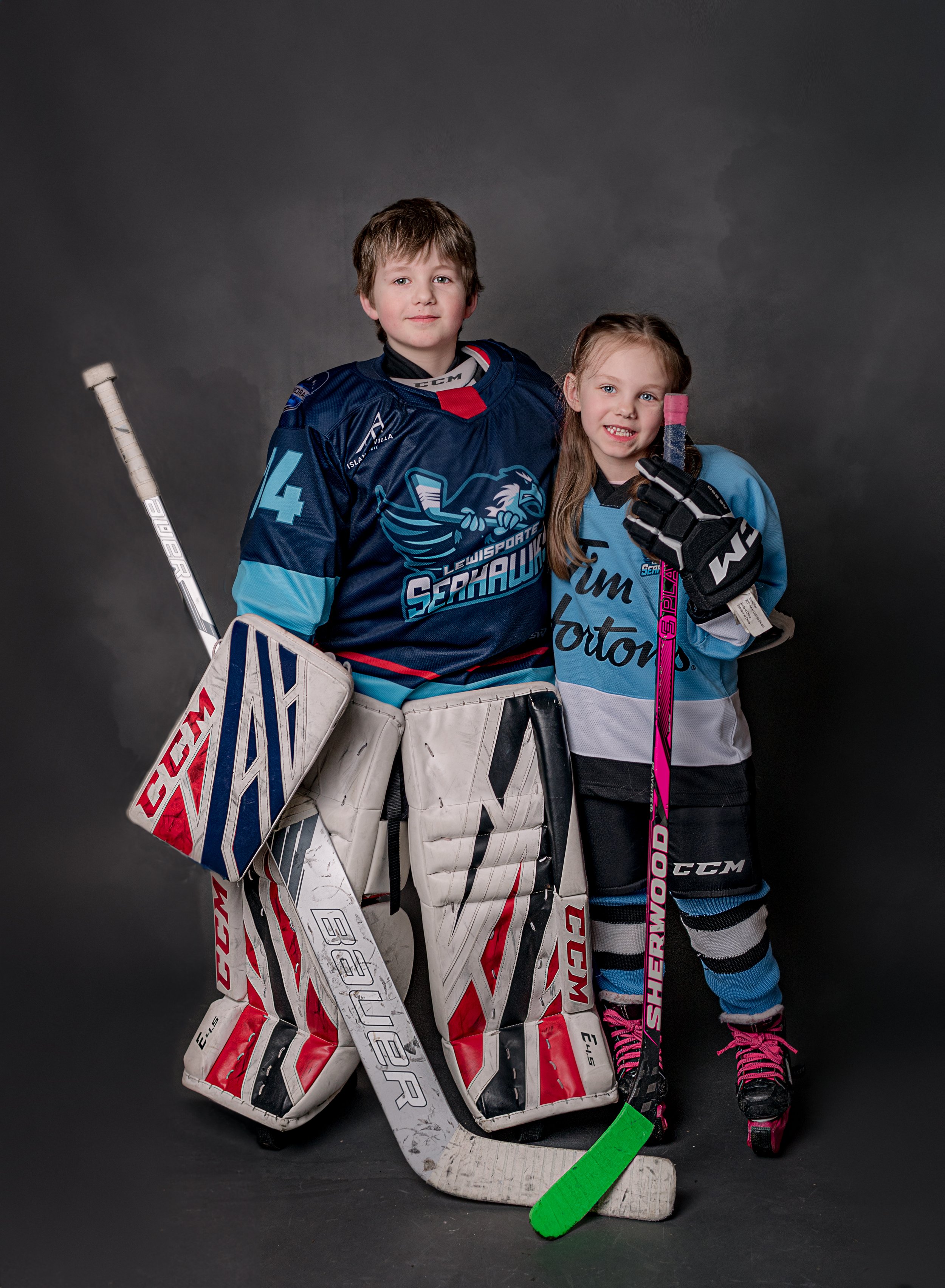 school age girl and boy stood up dressed in hockey equipment and hockey sticks for a portrait