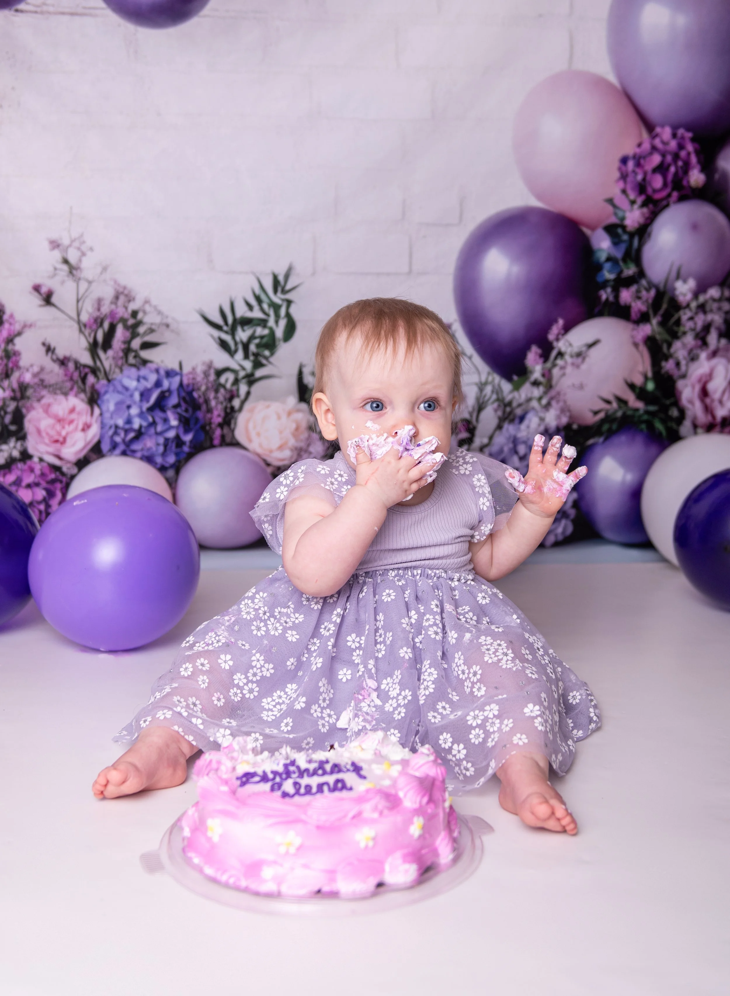 A young girl with blue eyes and light brown hair celebrating her birthday. She is sitting in front of a pink birthday cake decorated with flowers and written message, wearing a purple dress with floral patterns. She has cake and frosting on her face and hands, with purple and pink balloons and flowers in the background.