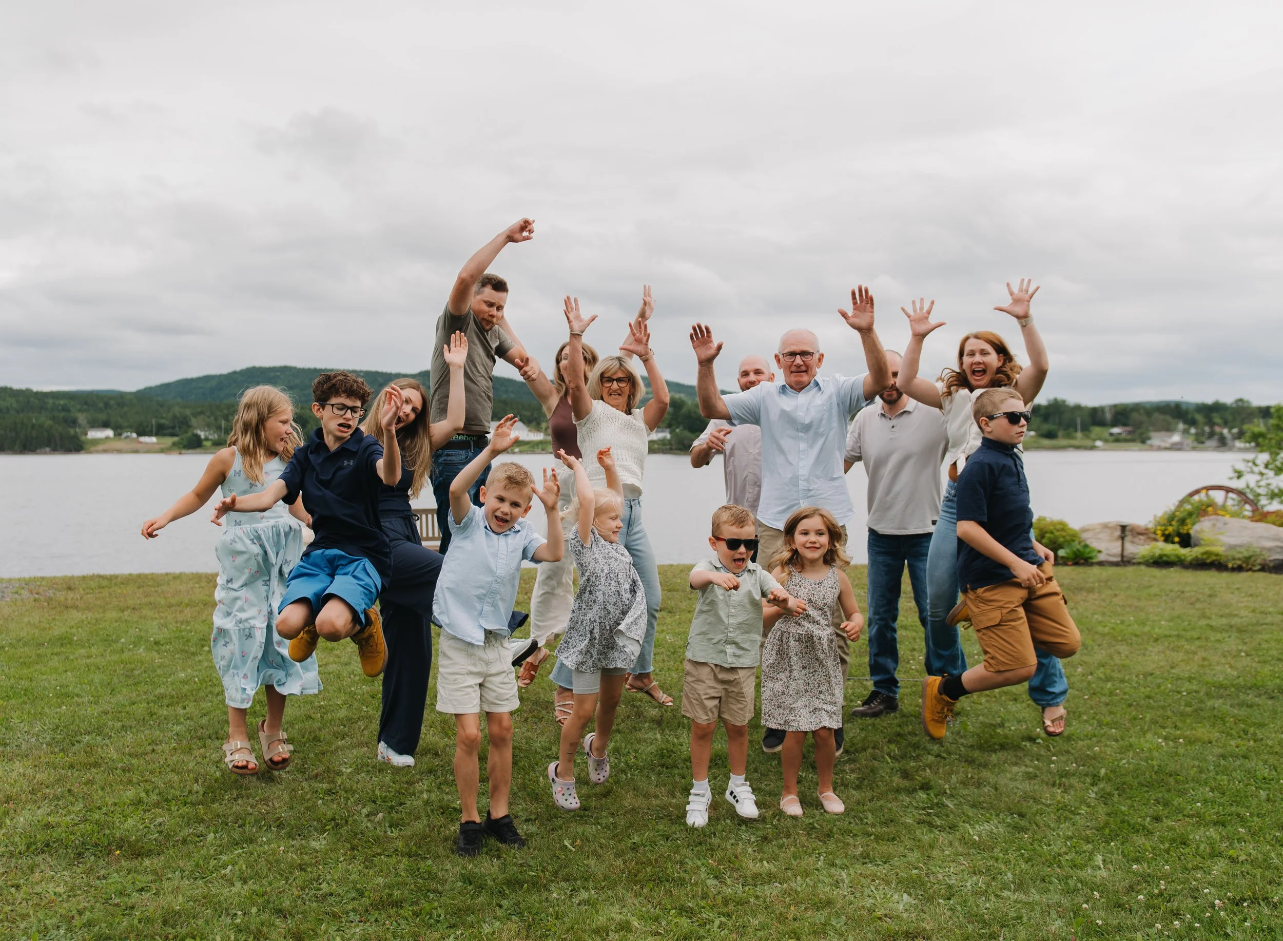 A large group of people, including children and adults, are outdoors on a grassy area near a body of water. They are dancing or celebrating with their hands raised, smiling, and appear to be happy.