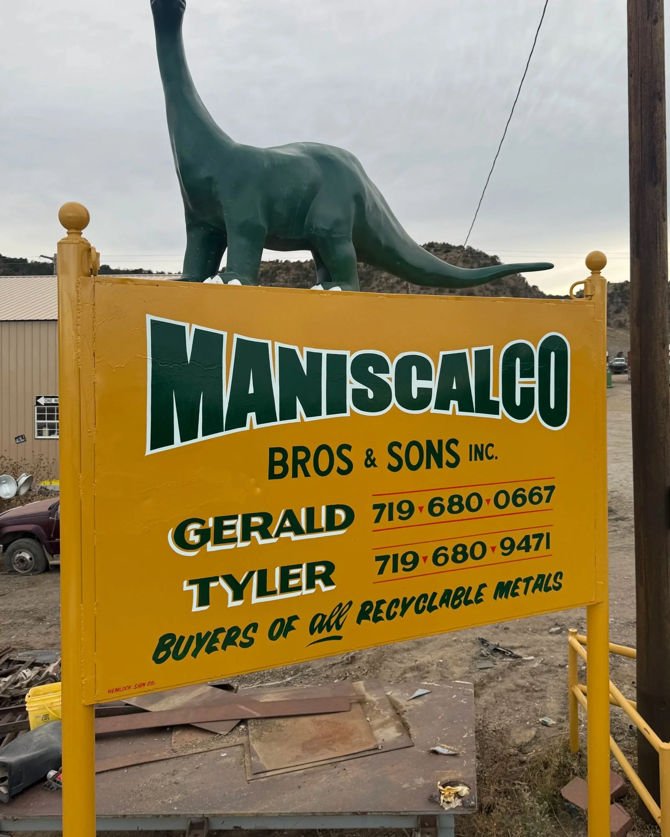 Yellow sign for Maniscalco Bros & Sons Inc. with phone numbers for Gerald and Tyler, topped with a large green dinosaur statue, outside on a cloudy day.
