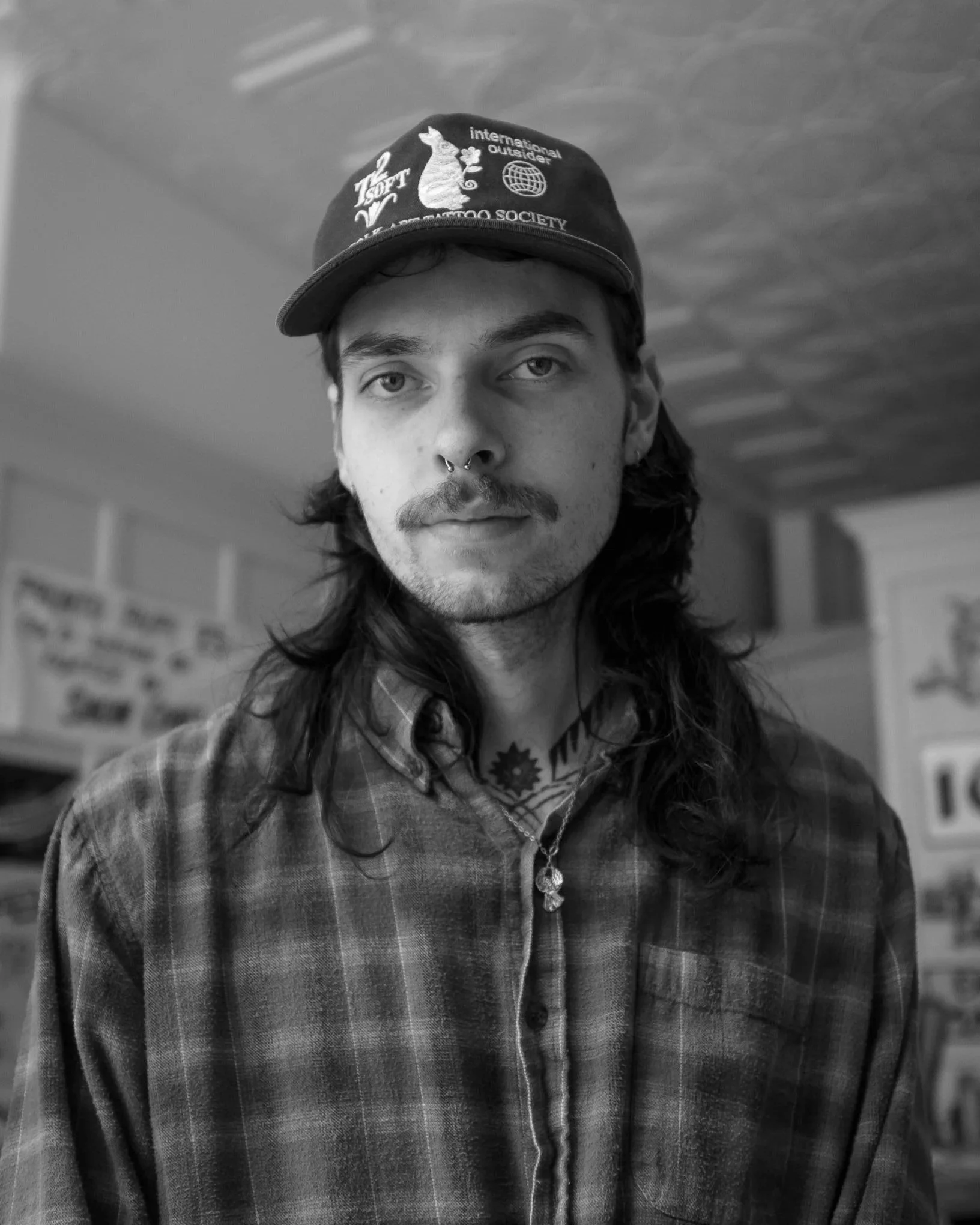 A young man with long hair, a mustache, and a beard, wearing a baseball cap, a plaid shirt, and a necklace, standing indoors with a blurred background.