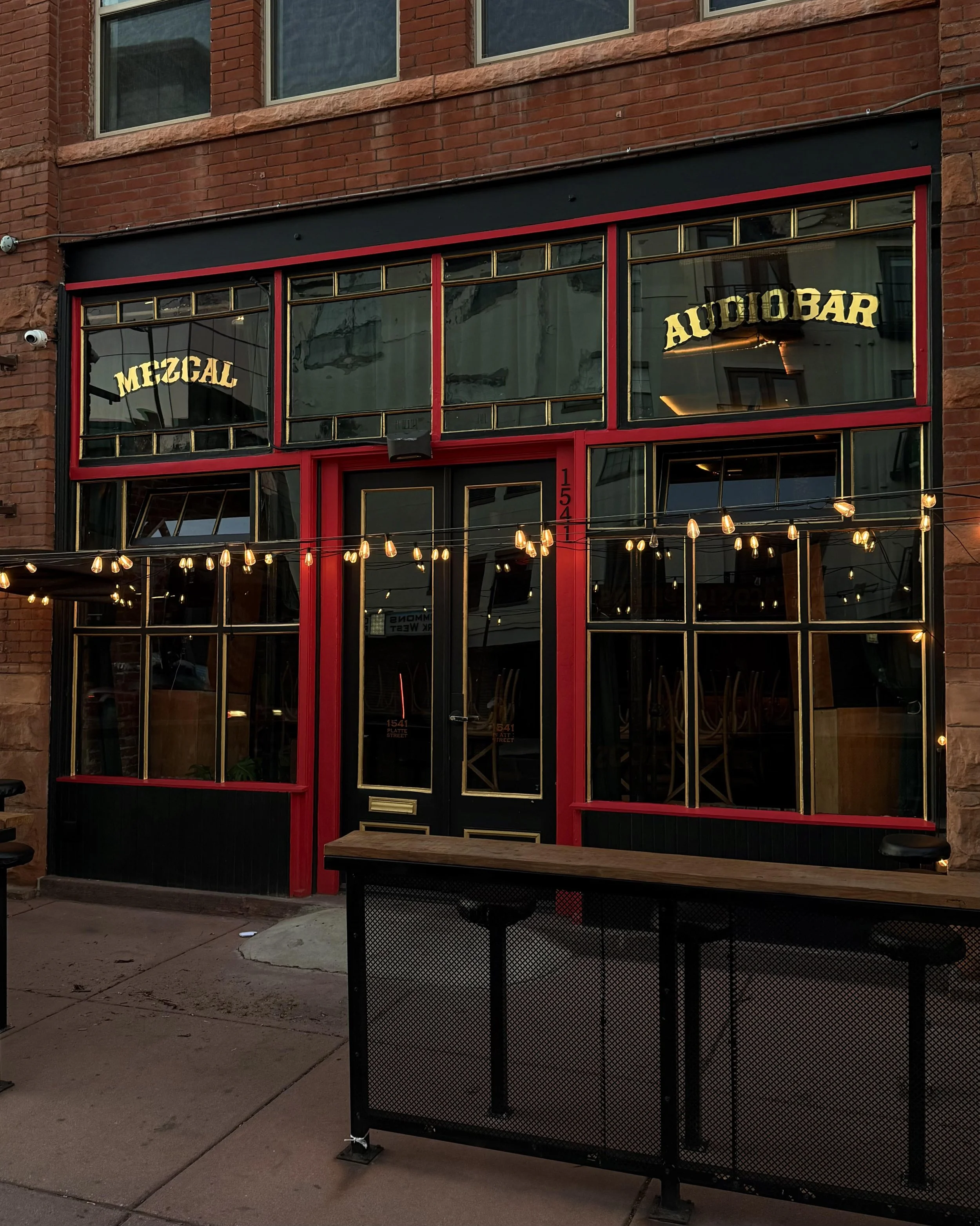 The exterior of a restaurant called 'Malinche Audiobar' in Denver with large windows, red framing, and signs inside reading 'Mezcal' and 'Audiobar'. There are string lights hanging outside and a black metal table with a wooden top in front.
