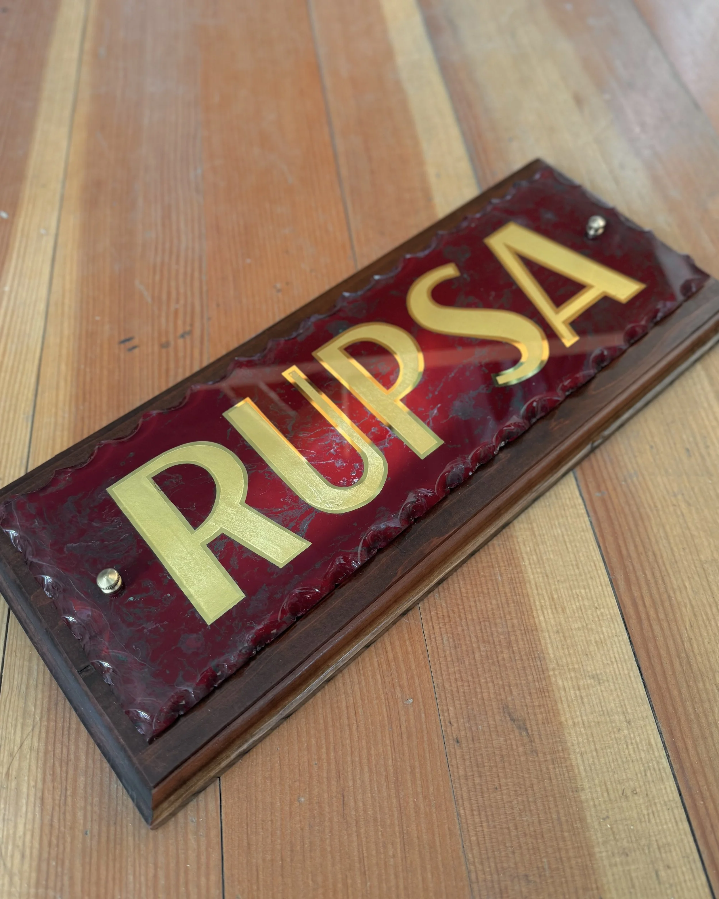 A rustic gold leaf and glass wooden sign with a red marbled glass background and gold lettering that says 'RUPSA', resting on a hardwood floor.